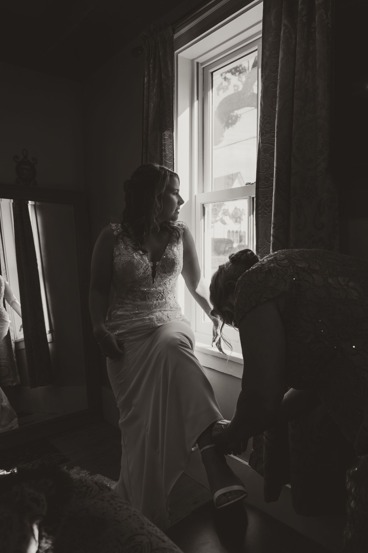 Black and white photo of a bride in lace gown seated by a window while someone helps with her shoes.