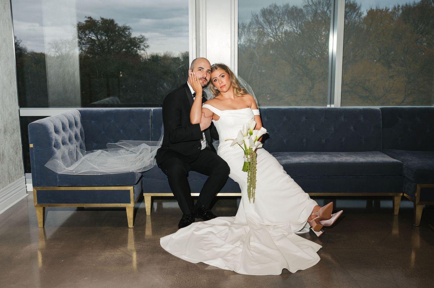 Bride and groom pose together on a blue velvet sofa in a modern venue with large windows.