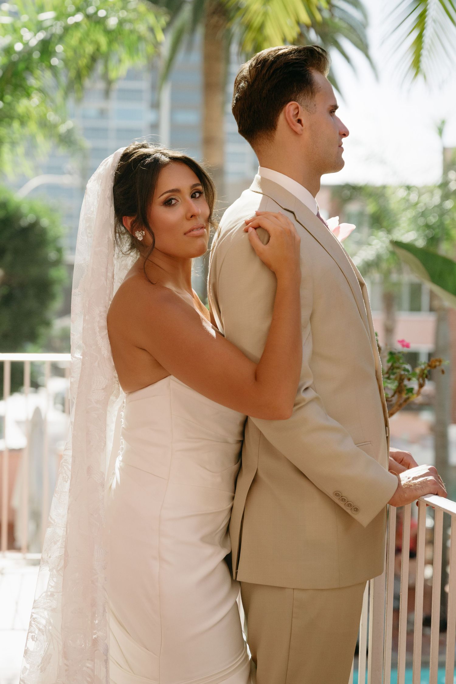 Bride in white gown and veil embraces groom in tan suit on a sunny balcony with palm trees in the background.