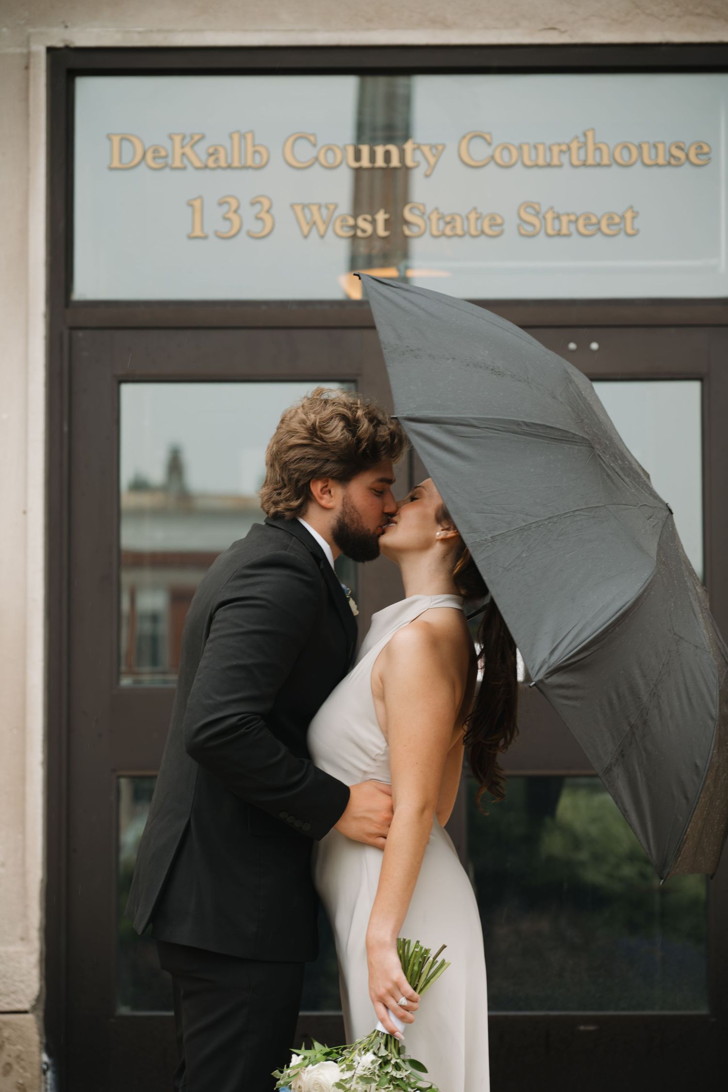 Couple kissing under umbrella outside DeKalb County Courthouse on their wedding day.