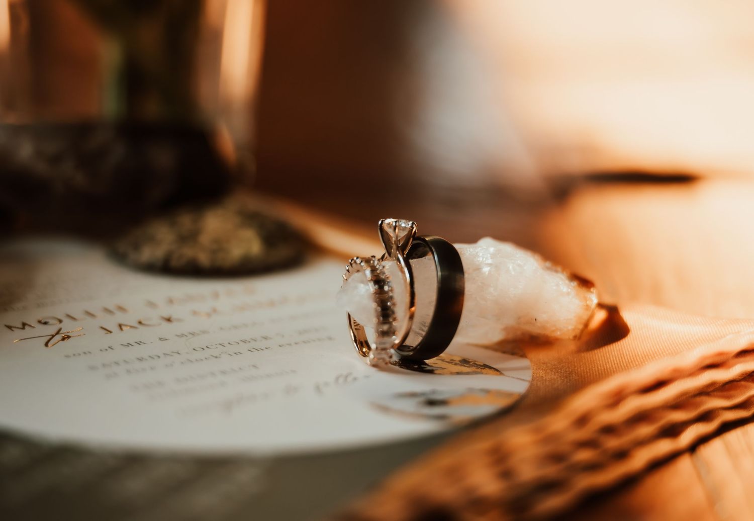 Close-up of wedding rings and engagement ring resting on wedding invitation with warm golden light.