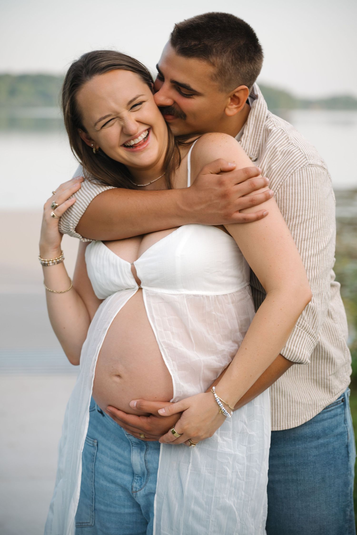 Happy pregnant woman in white dress embraced by her partner outdoors near a lake, both smiling joyfully.