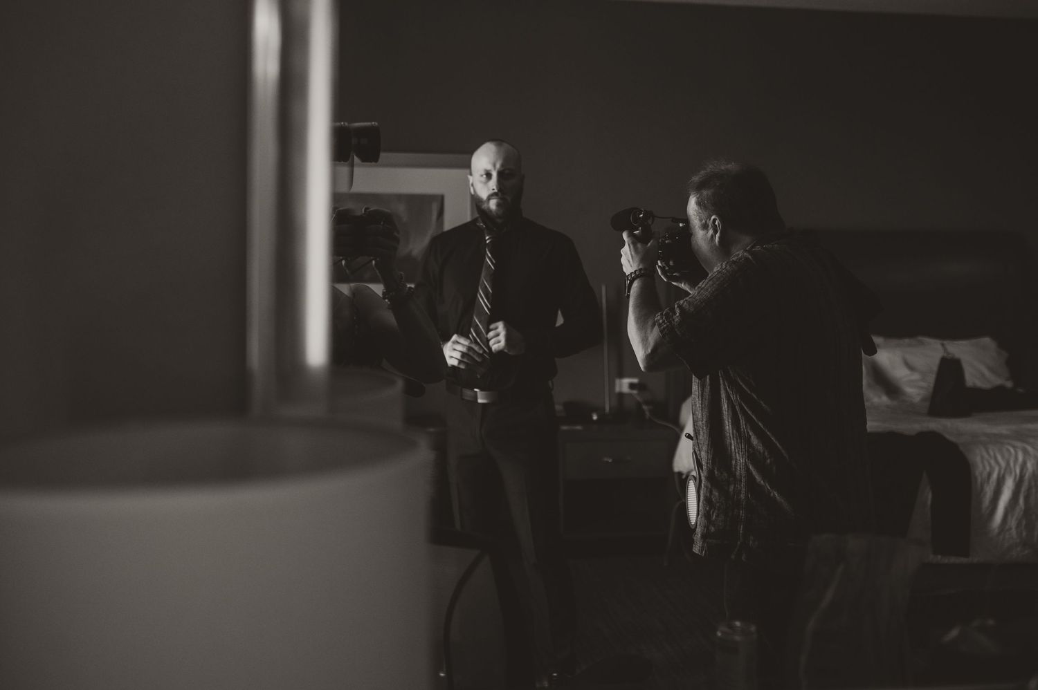 Black and white photo of groom being photographed by videographer while getting ready in hotel room.