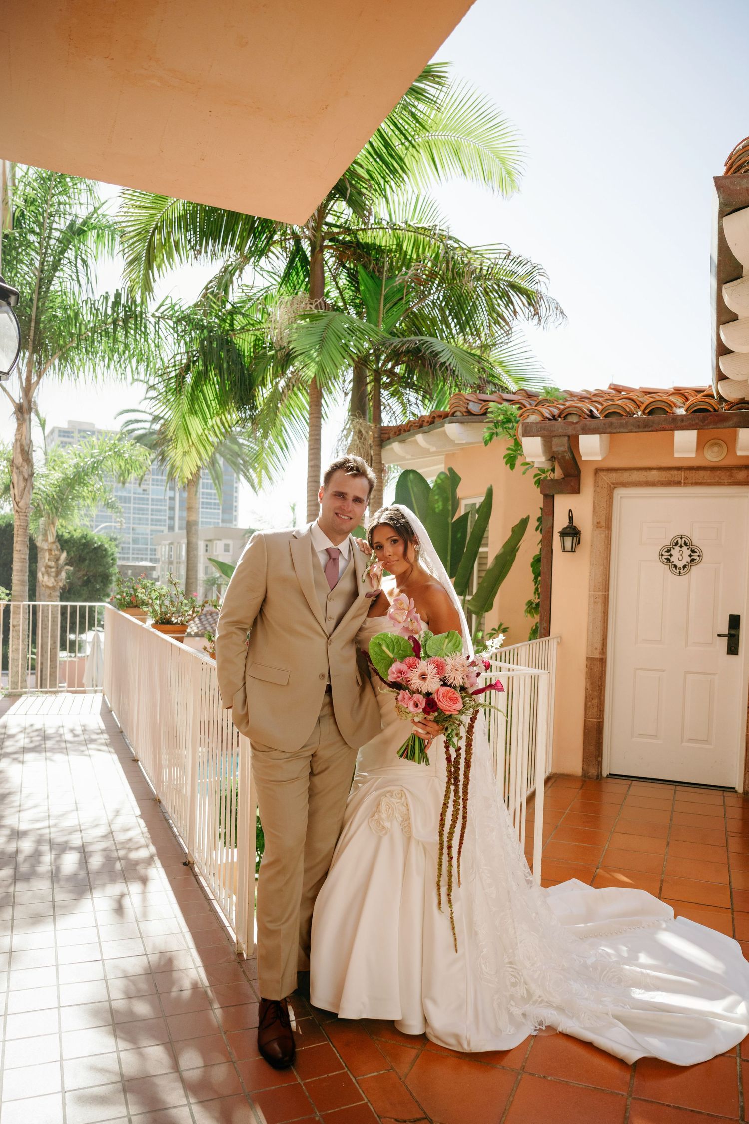 Bride and groom pose on a sunlit terrace with palm trees, she holds a pink bouquet in her white gown, he wears a tan suit. California.