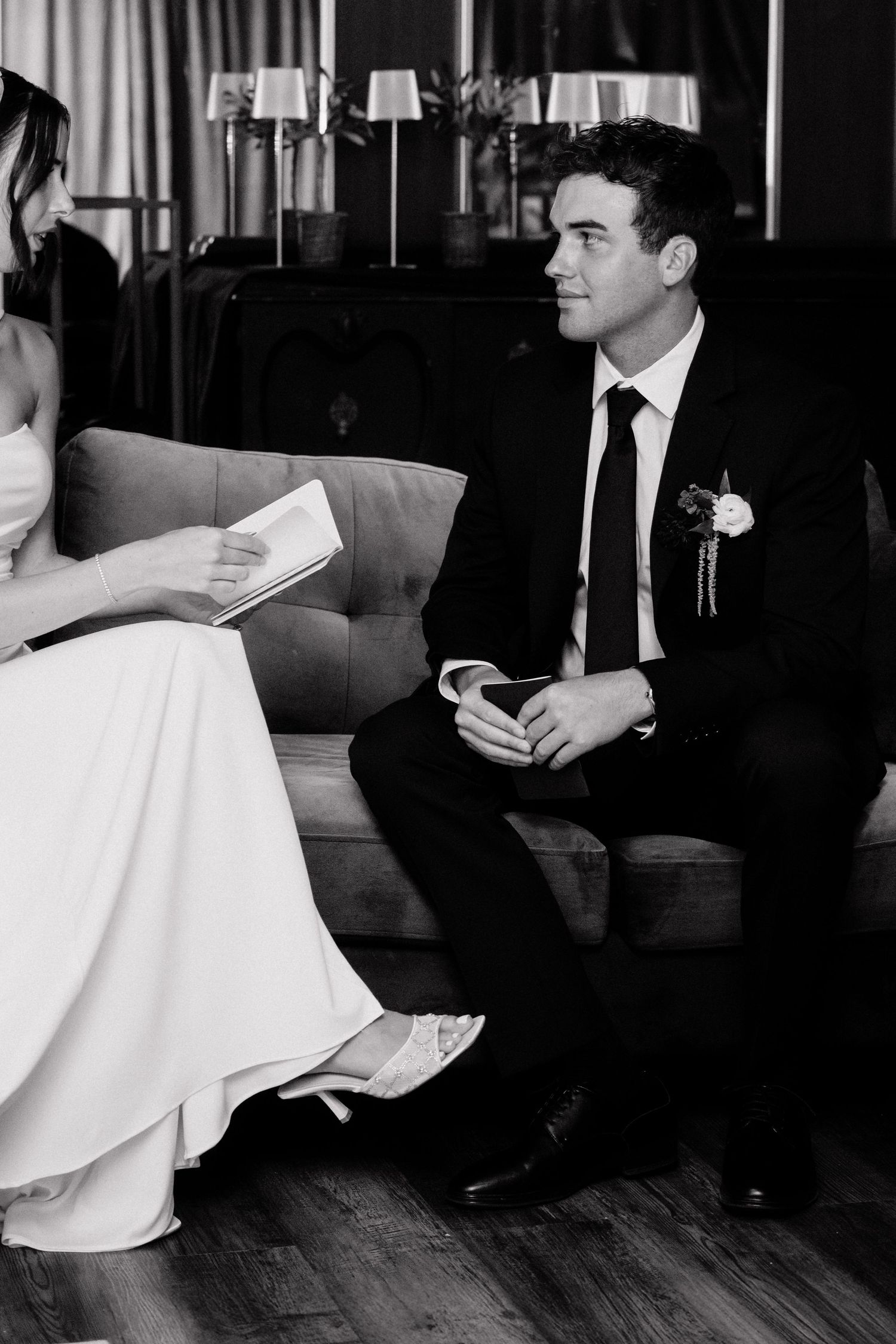 Black and white wedding photo of groom in suit sitting on sofa listening to bride read vows from a book.