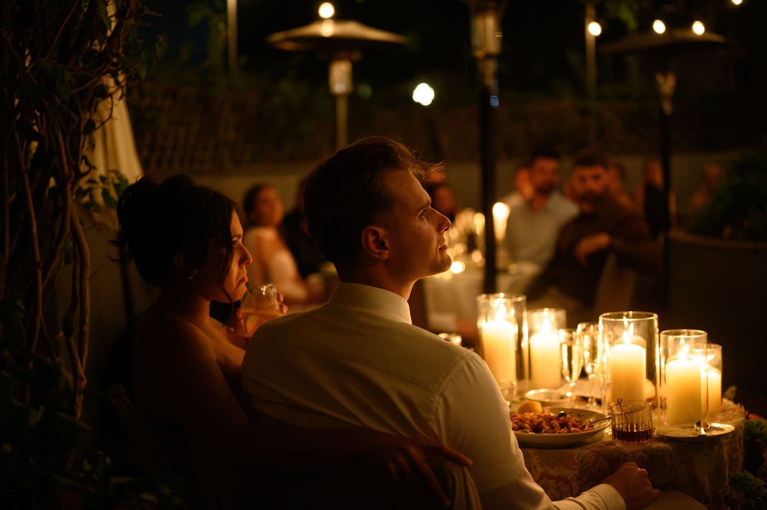 Couple sitting together at a candlelit outdoor evening wedding reception with glowing candles on the table.