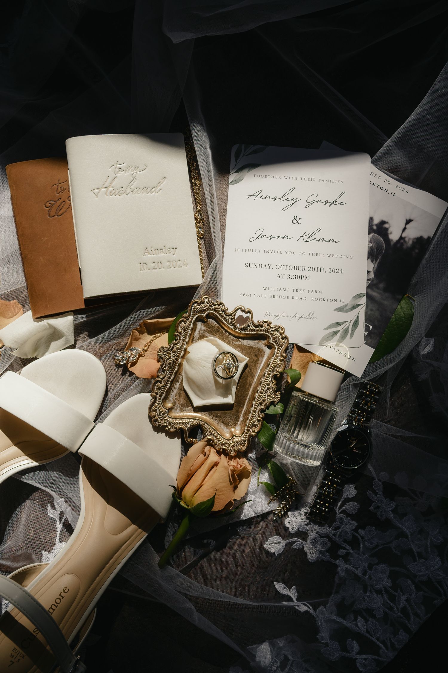 Elegant wedding flat lay with invitations, rings, white heels, perfume, and floral details on dark fabric.