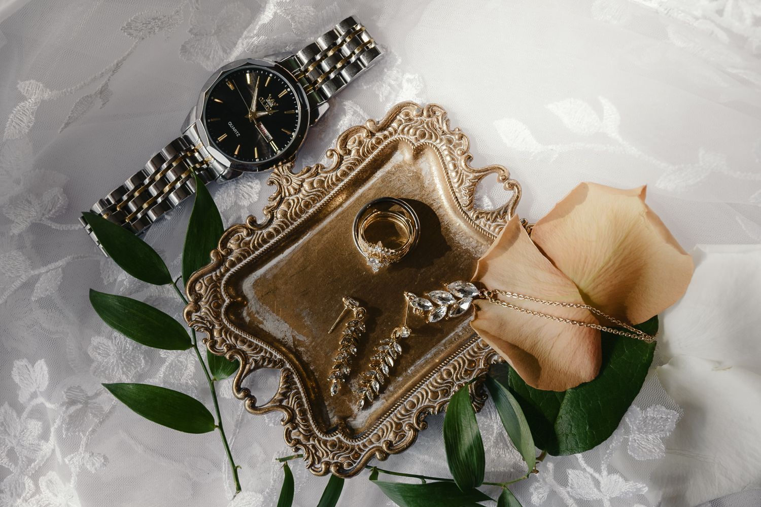 Wedding flat lay featuring gold ring dish, silver watch, wedding band, and peach rose on white lace.