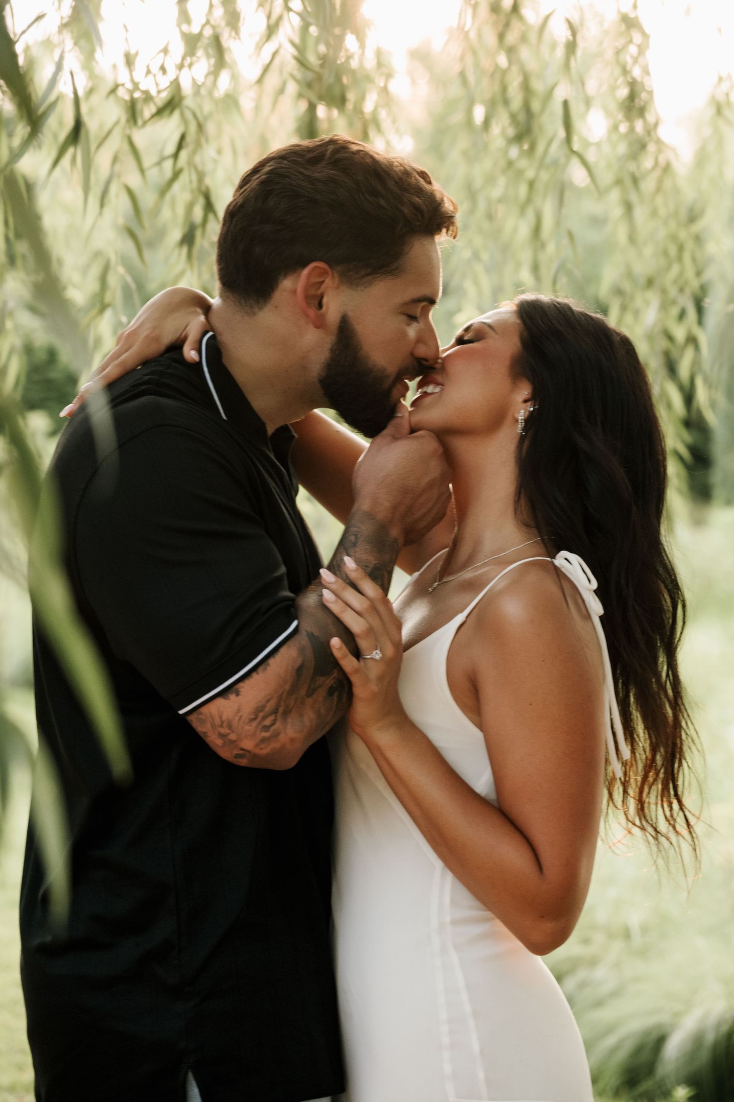 Couple shares a romantic almost-kiss embrace surrounded by lush green weeping willow branches at sunset.