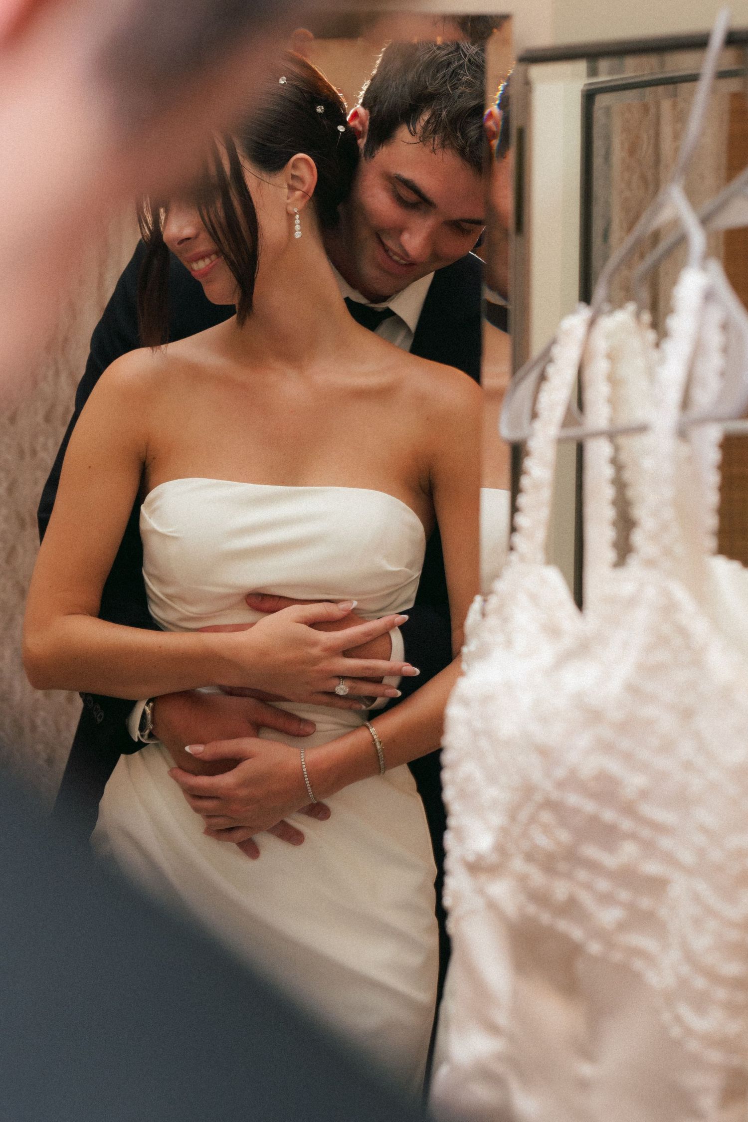 A groom tenderly embraces his bride in a white strapless wedding dress, smiling warmly near hanging bridal gowns.
