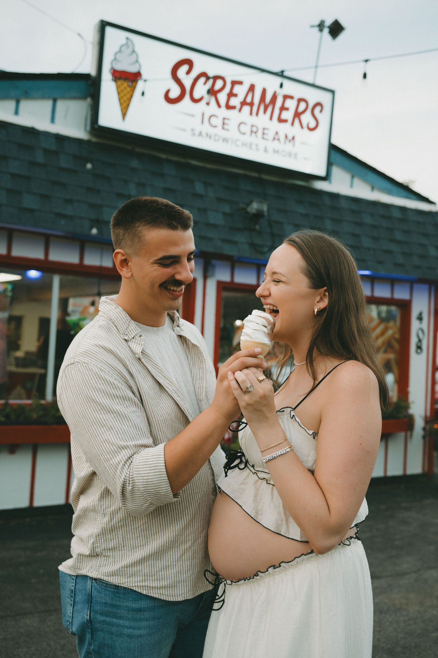 Pregnant couple sharing ice cream cone outside Screamers Ice Cream shop, smiling and enjoying a sweet moment together.