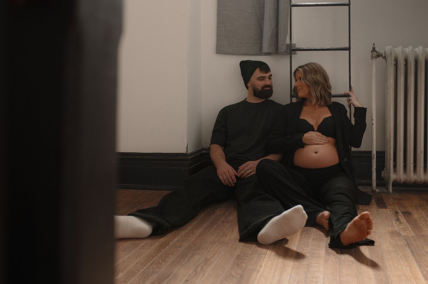 Expecting couple dressed in black sitting together on a wooden floor, sharing a quiet intimate moment indoors.