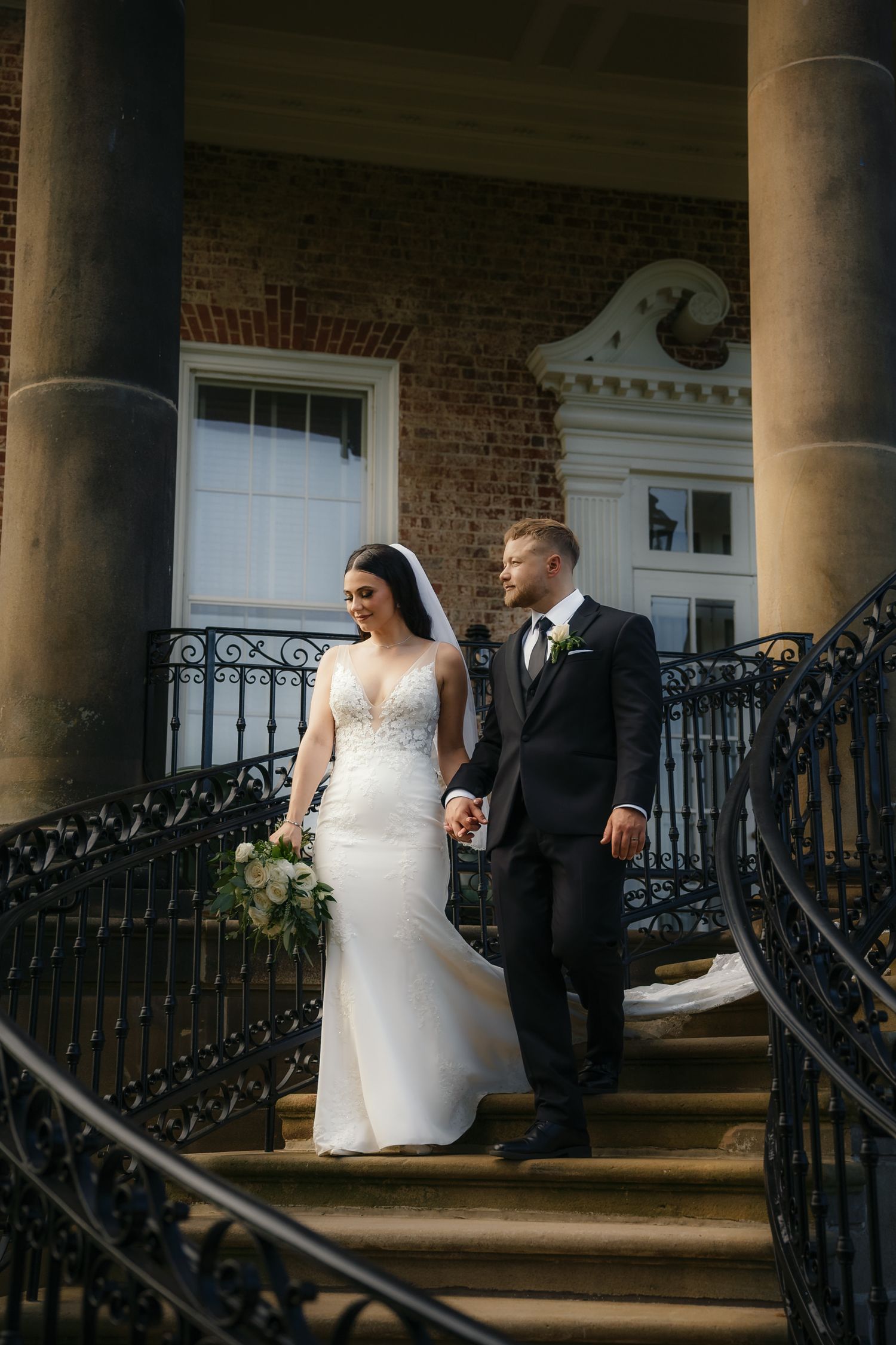Bride and groom holding hands descending elegant staircase of historic brick mansion with iron railings.