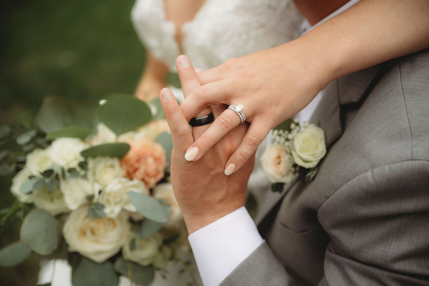 Close-up of bride and groom holding hands showing diamond engagement ring with a floral bouquet.