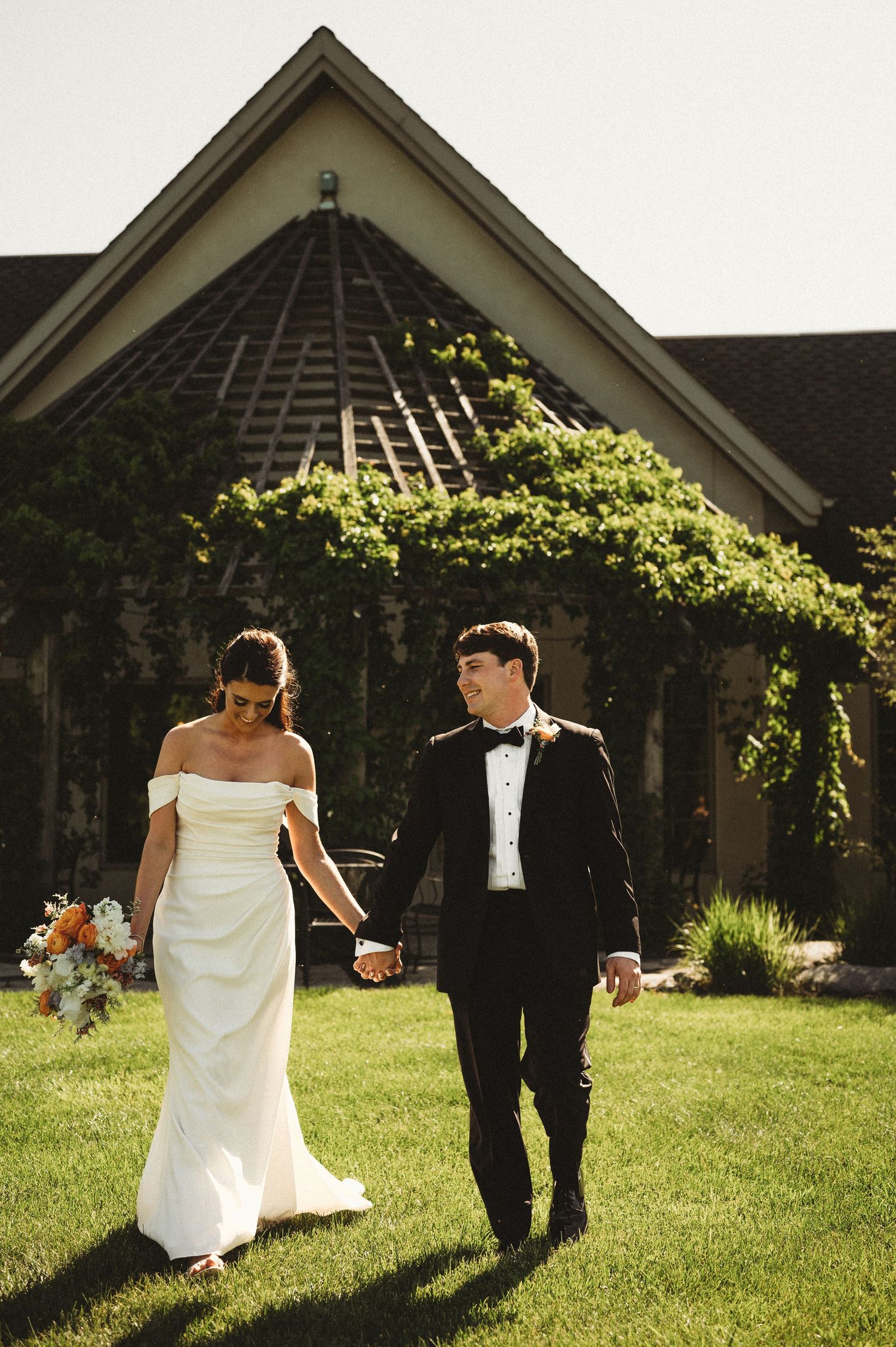 Bride and groom walk hand in hand on green lawn in front of an ivy-covered wedding venue.