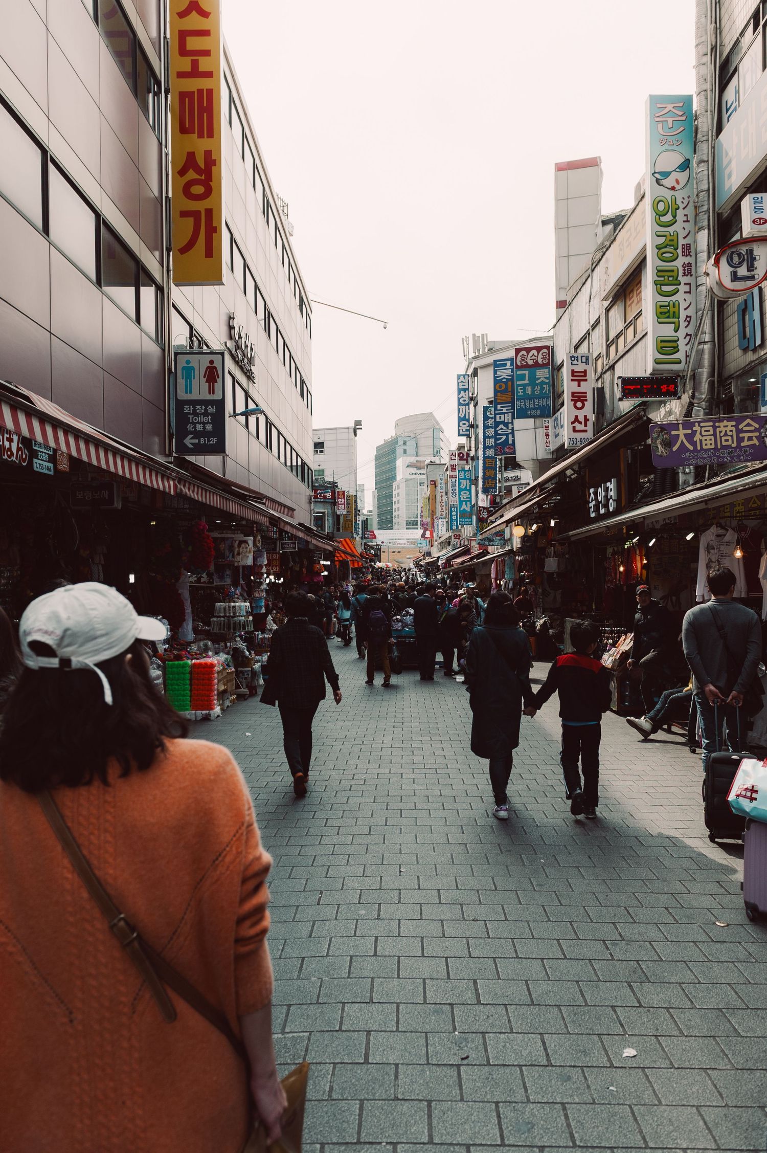 Pedestrians walking through a busy shopping street in South Korea, surrounded by storefronts and colorful Korean signs.