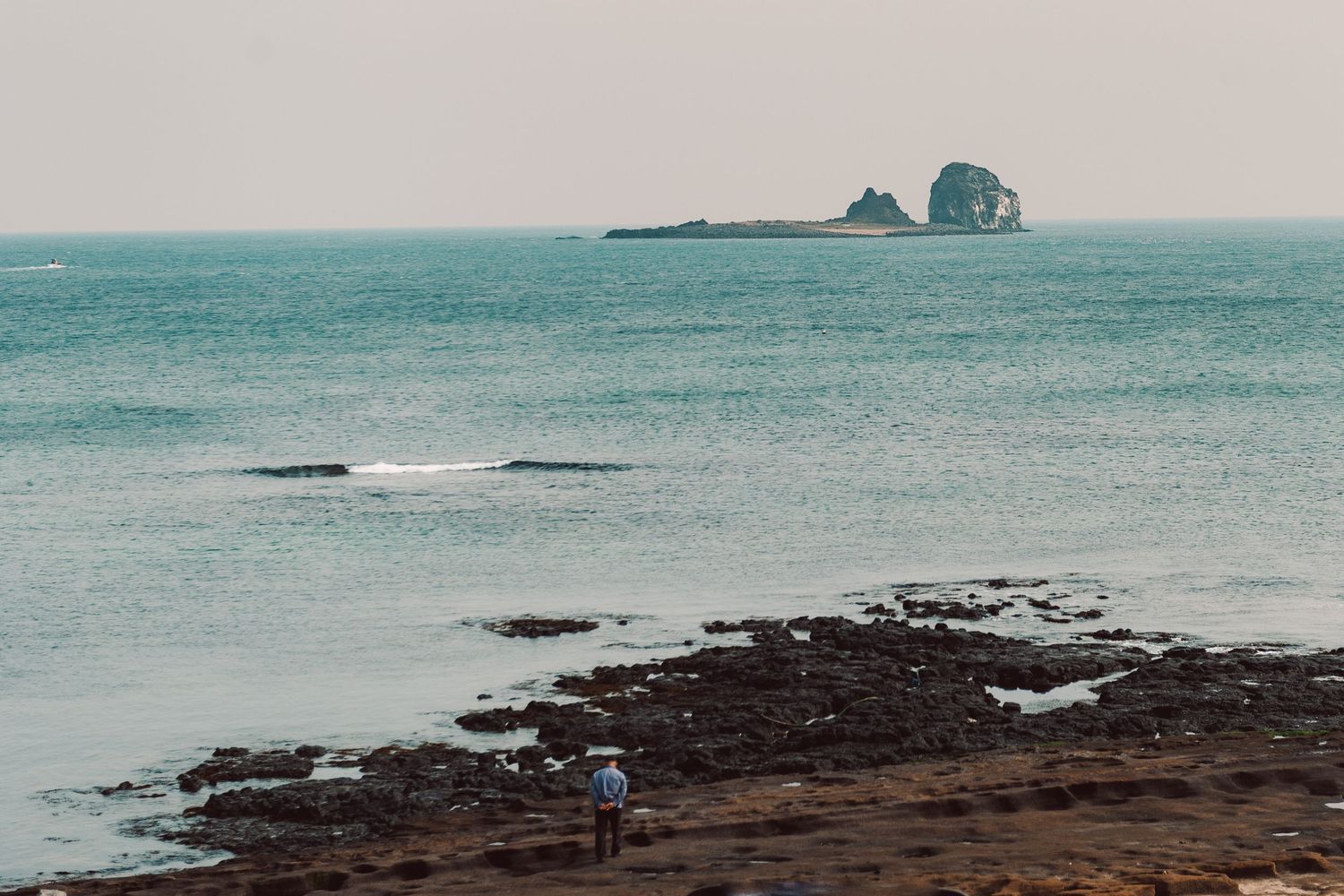 Minimal coastal landscape with calm turquoise water, a distant rocky island, and a pale overcast sky off of Jeju Island, South Korea.