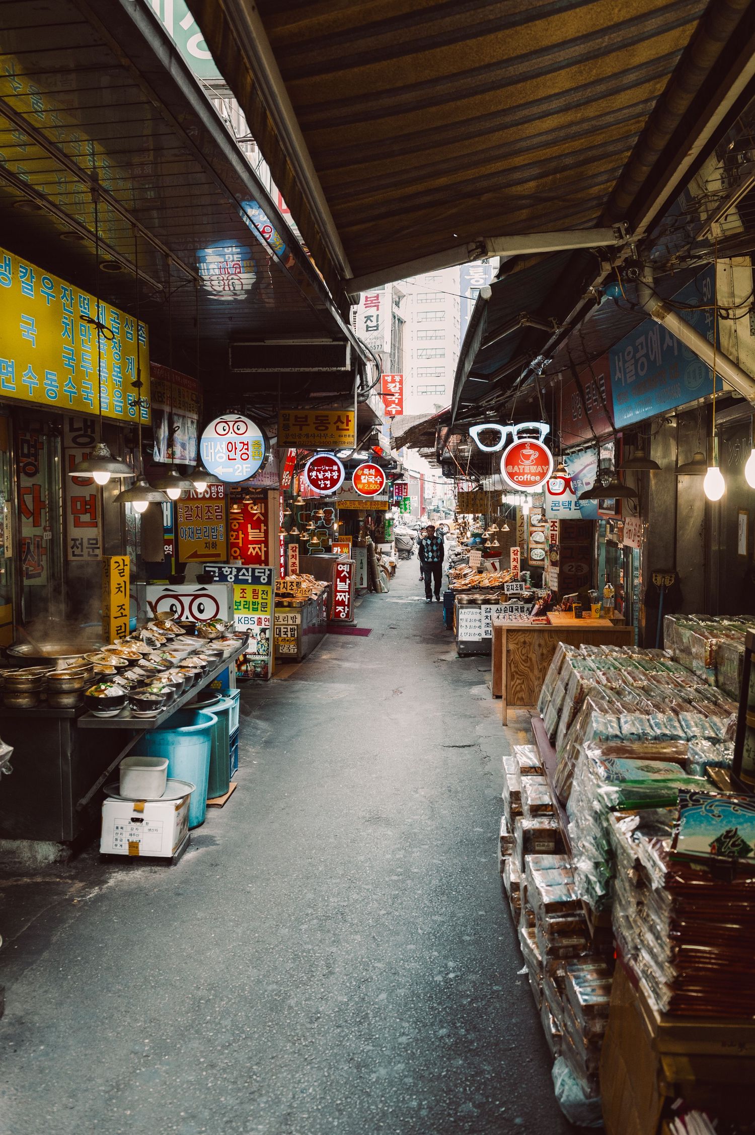 Narrow aisle inside a traditional Korean market, with food stalls, hanging signs, and vendors beneath a covered roof.  Seoul, South Korea.