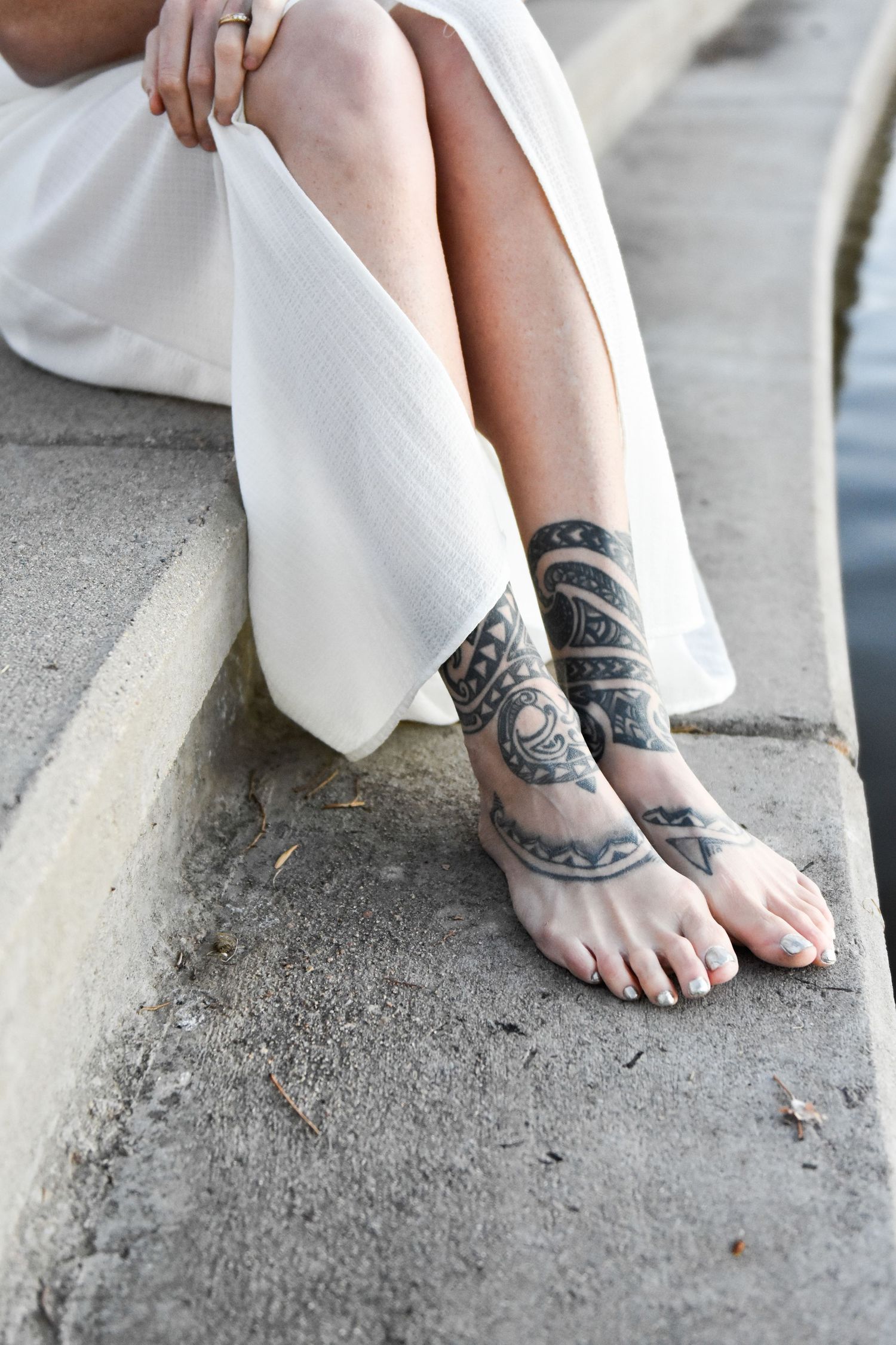 Close-up of woman's tattooed feet and ankles with tribal designs, wearing a white dress, seated on stone steps.