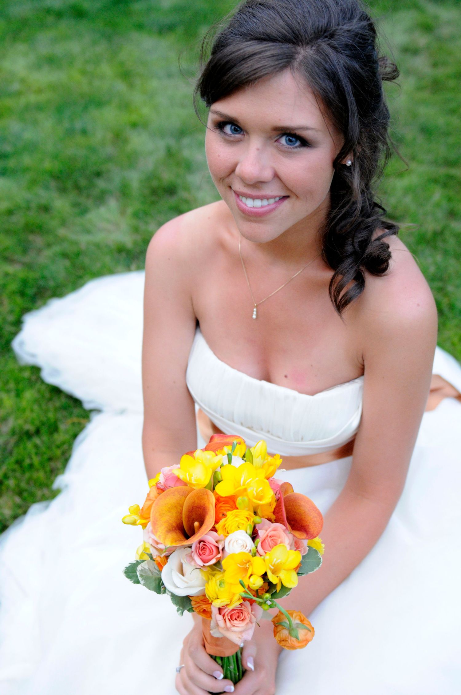 Smiling bride in white strapless gown holding a vibrant yellow and orange wedding bouquet on grass.