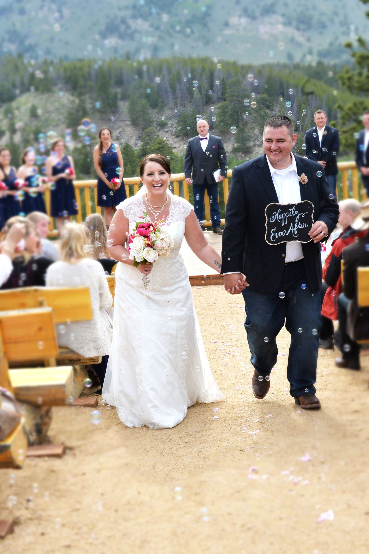 Newlywed couple walks down the aisle holding hands amid bubbles at an outdoor mountain wedding ceremony.