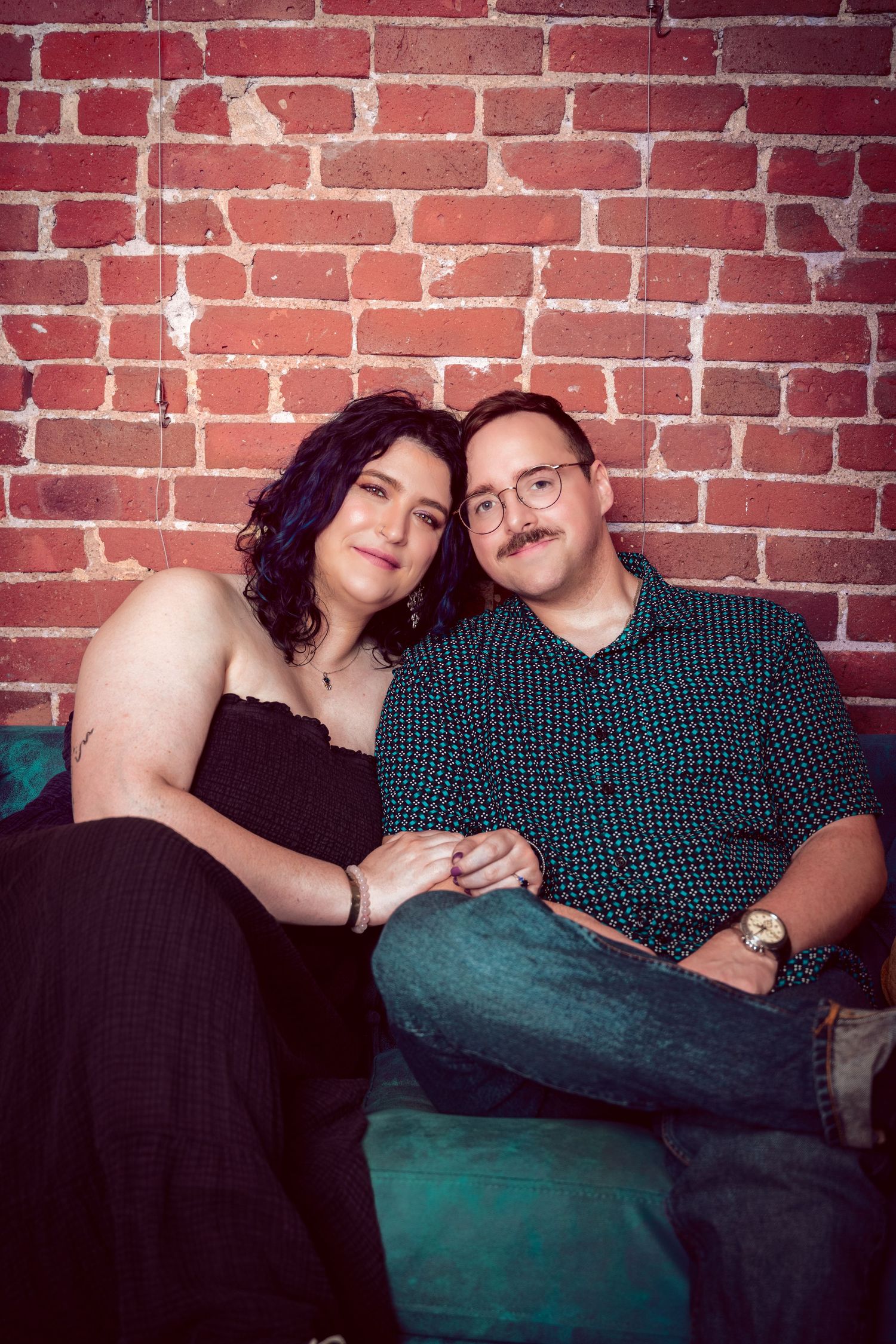 Couple smiling and holding hands while seated together against a red brick wall indoors.