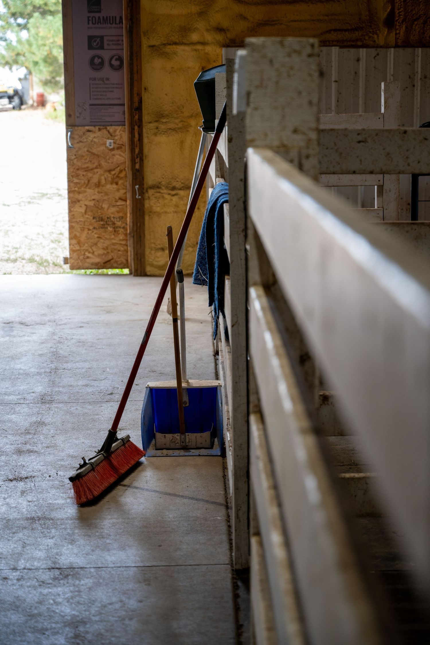 Red broom and dustpan leaning against wooden stable wall in a barn aisle with concrete floor.
