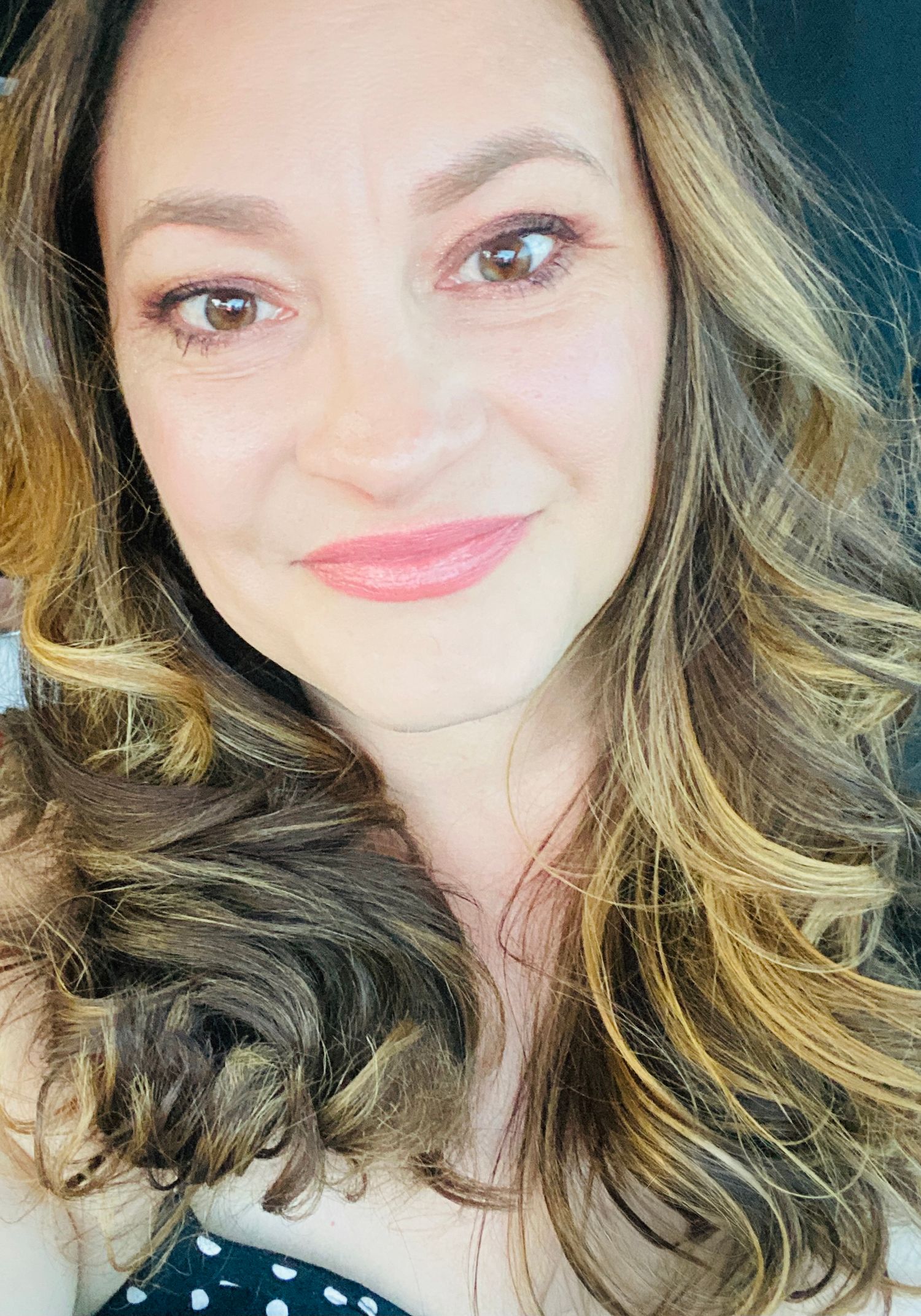 Smiling woman with wavy blonde highlighted hair holding a small dog in a selfie.