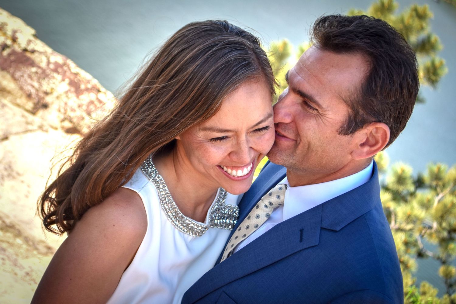 Happy bride smiling as groom in blue suit whispers in her ear during outdoor wedding.