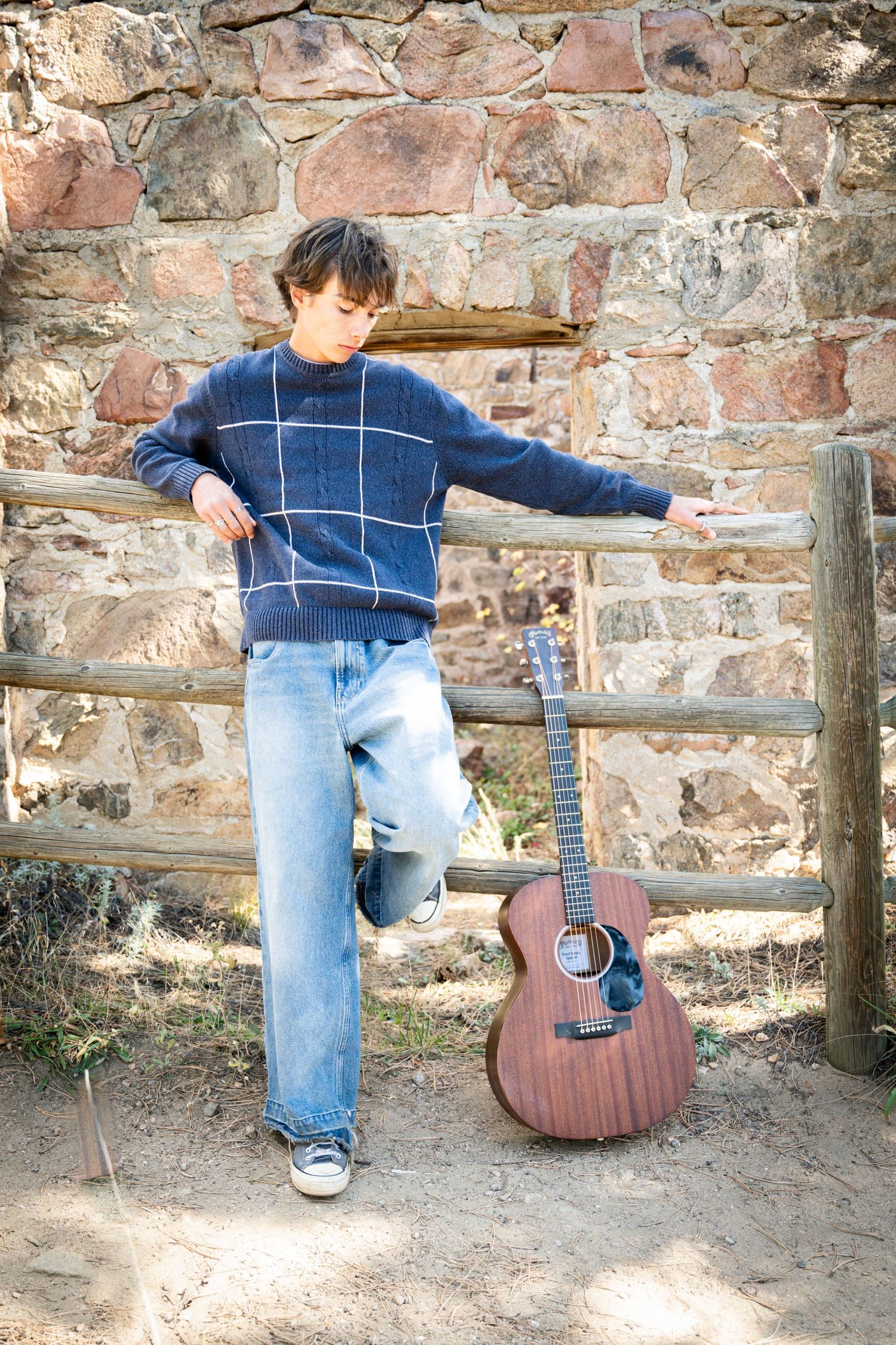 Teen boy in plaid sweater leaning on wooden fence beside acoustic guitar and stone wall.