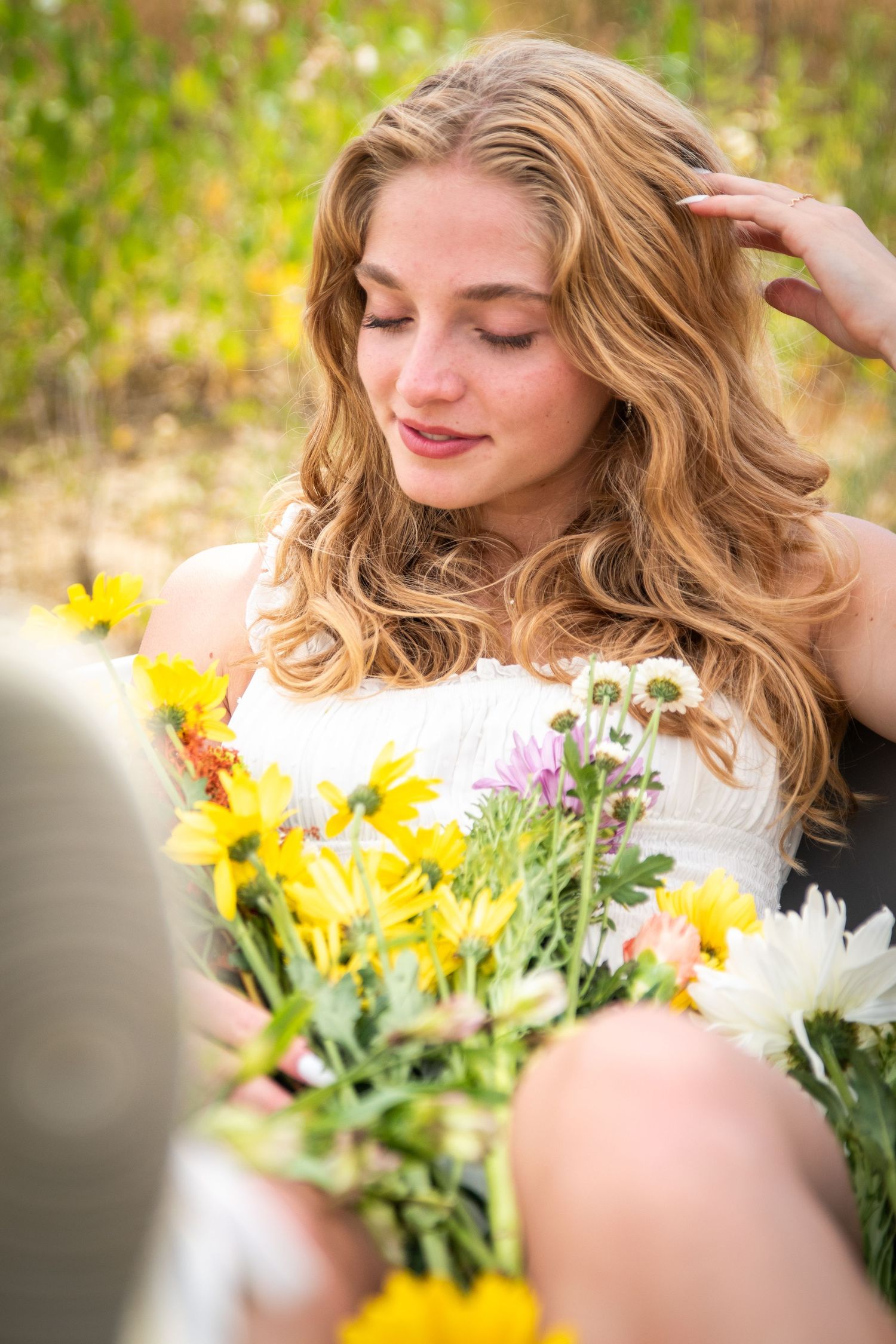 Young blonde woman with wavy hair sits in a field holding a colorful bouquet of wildflowers.