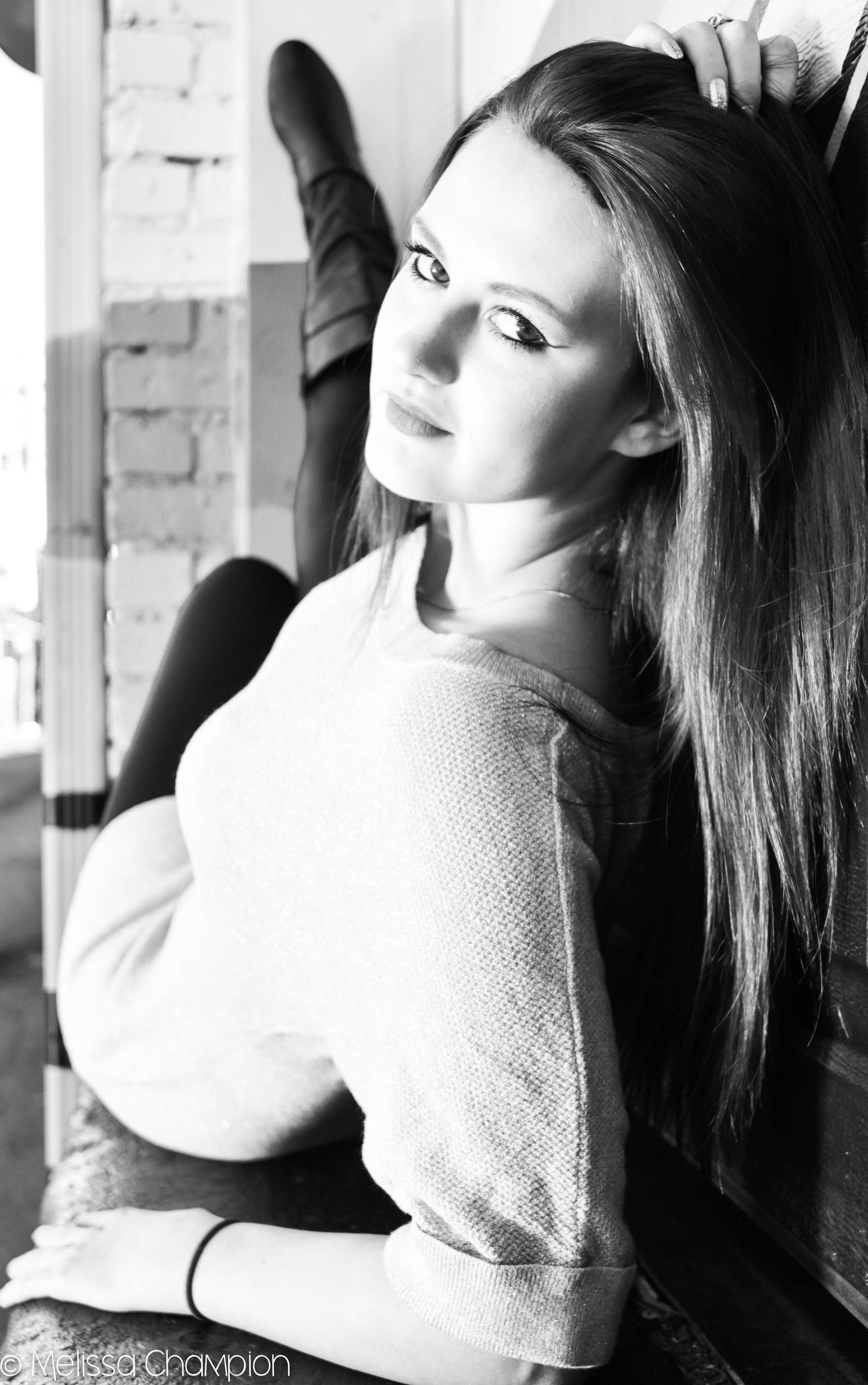 Black and white portrait of a young woman with long hair in a white sweater, posing thoughtfully against a brick wall.