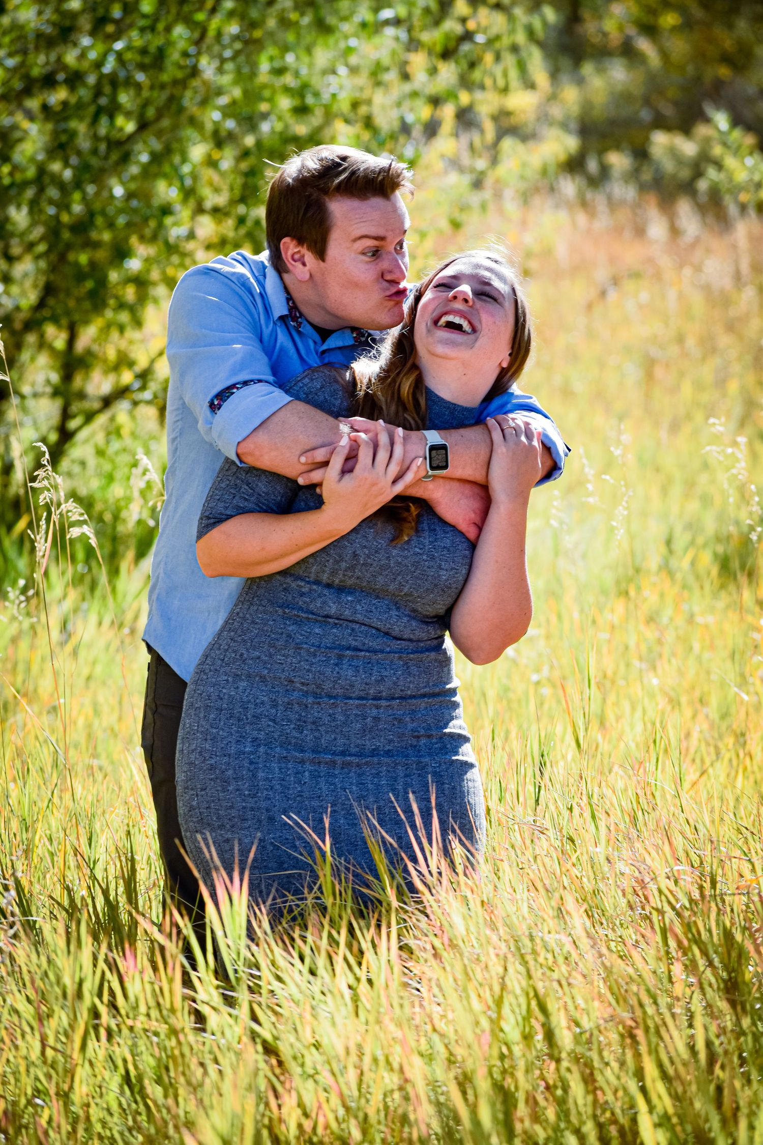 Couple laughing outdoors in tall grass as man in blue shirt hugs woman from behind.