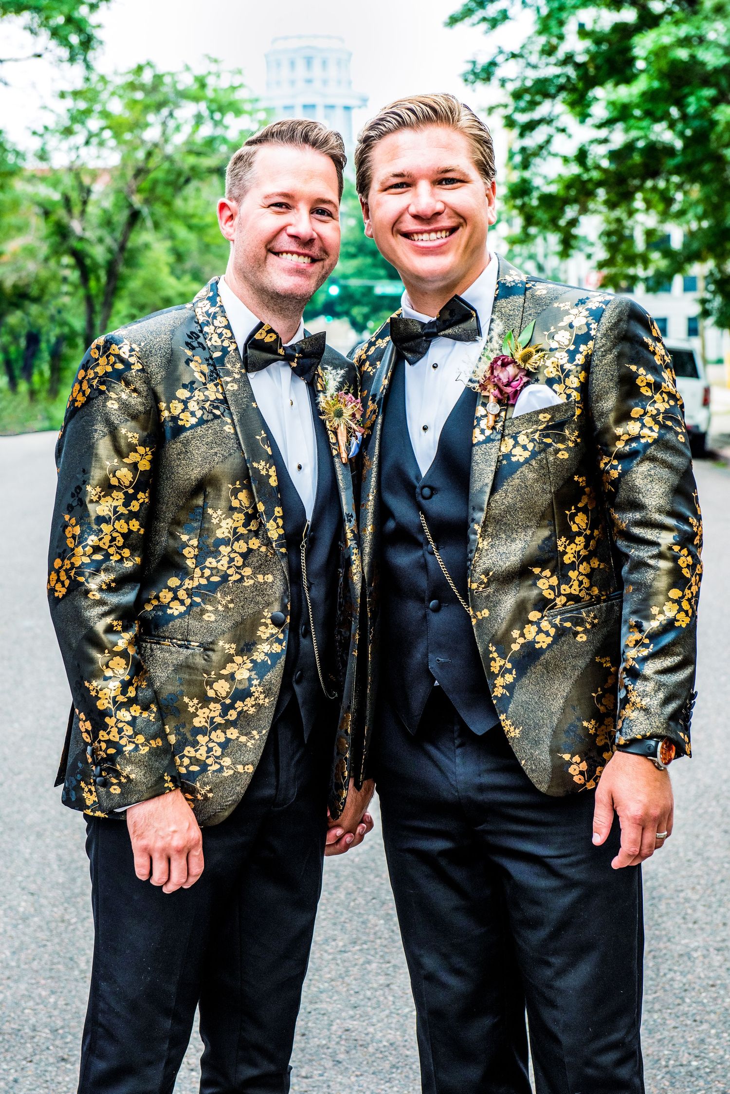 Two grooms in matching black and gold floral tuxedo jackets with bow ties smile together on their wedding day.