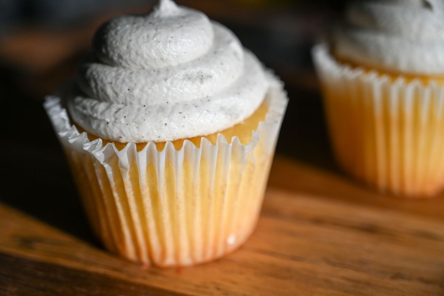 Close-up of a vanilla cupcake with gray swirled frosting in a white paper liner on wood.