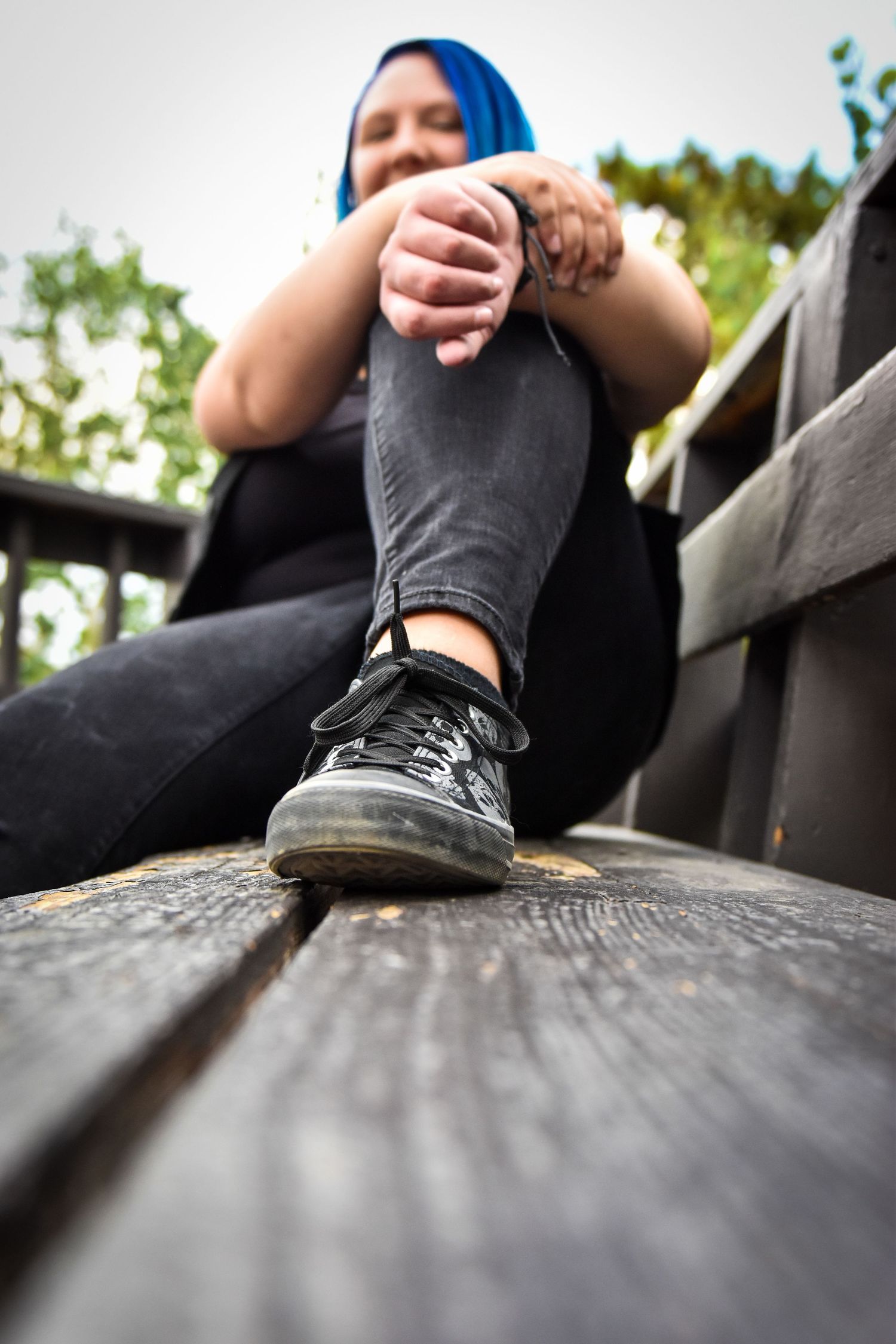 Young woman with blue hair sitting on wooden bench outdoors, wearing black jeans and sneakers.