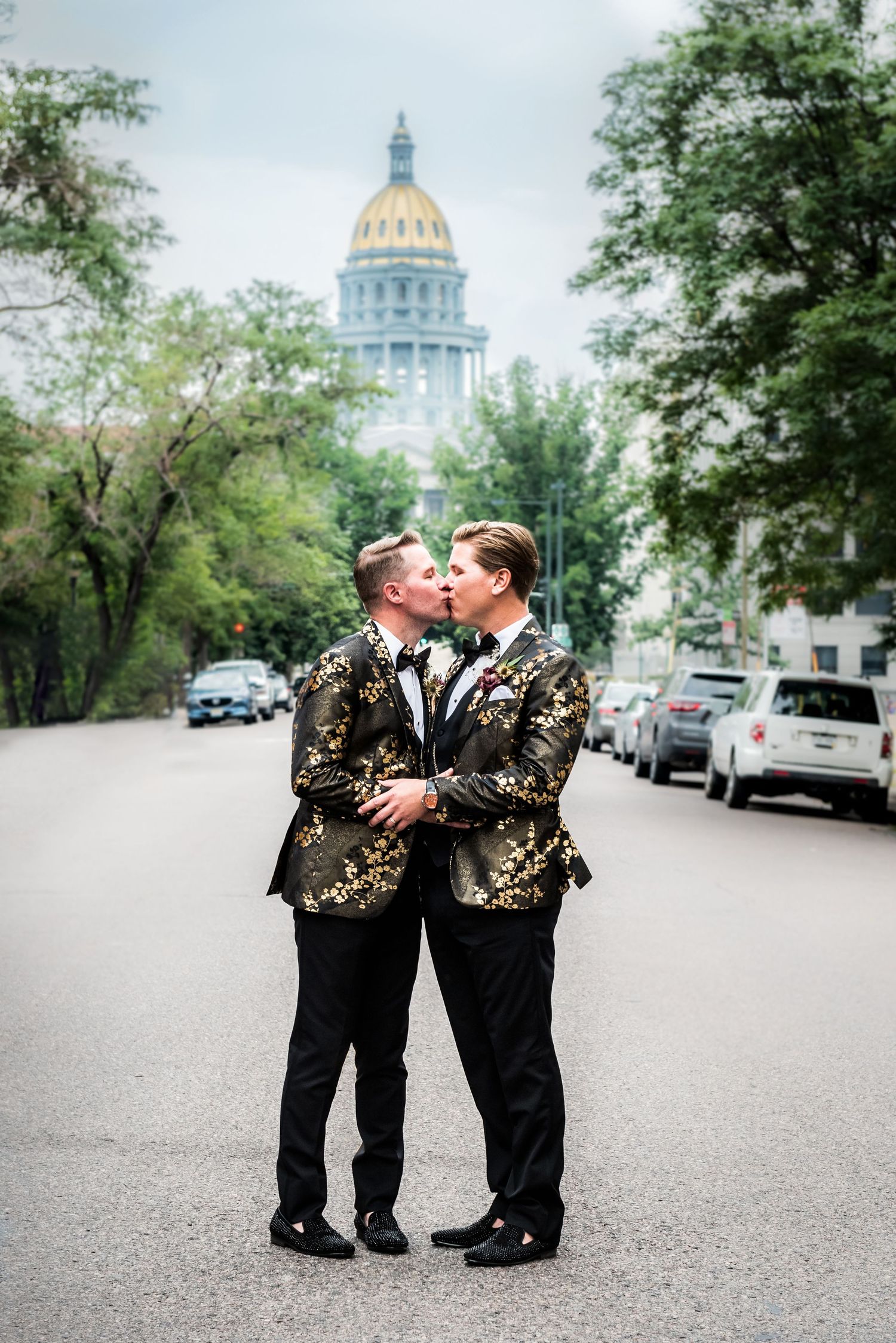 Two grooms kiss in matching black and gold floral tuxedos on street with Colorado Capitol dome backdrop.