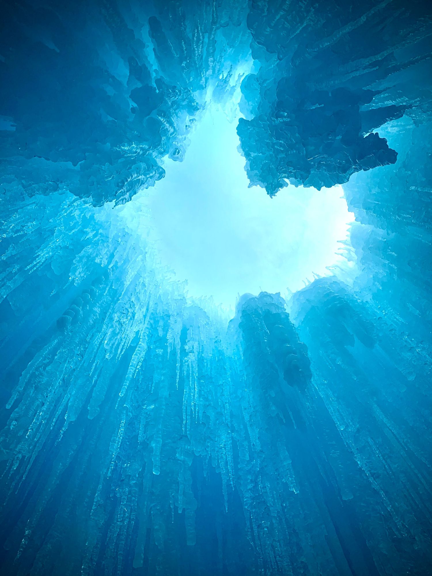 Stunning underwater view looking up through a heart-shaped hole in blue ice cave with icicles and light streaming through.