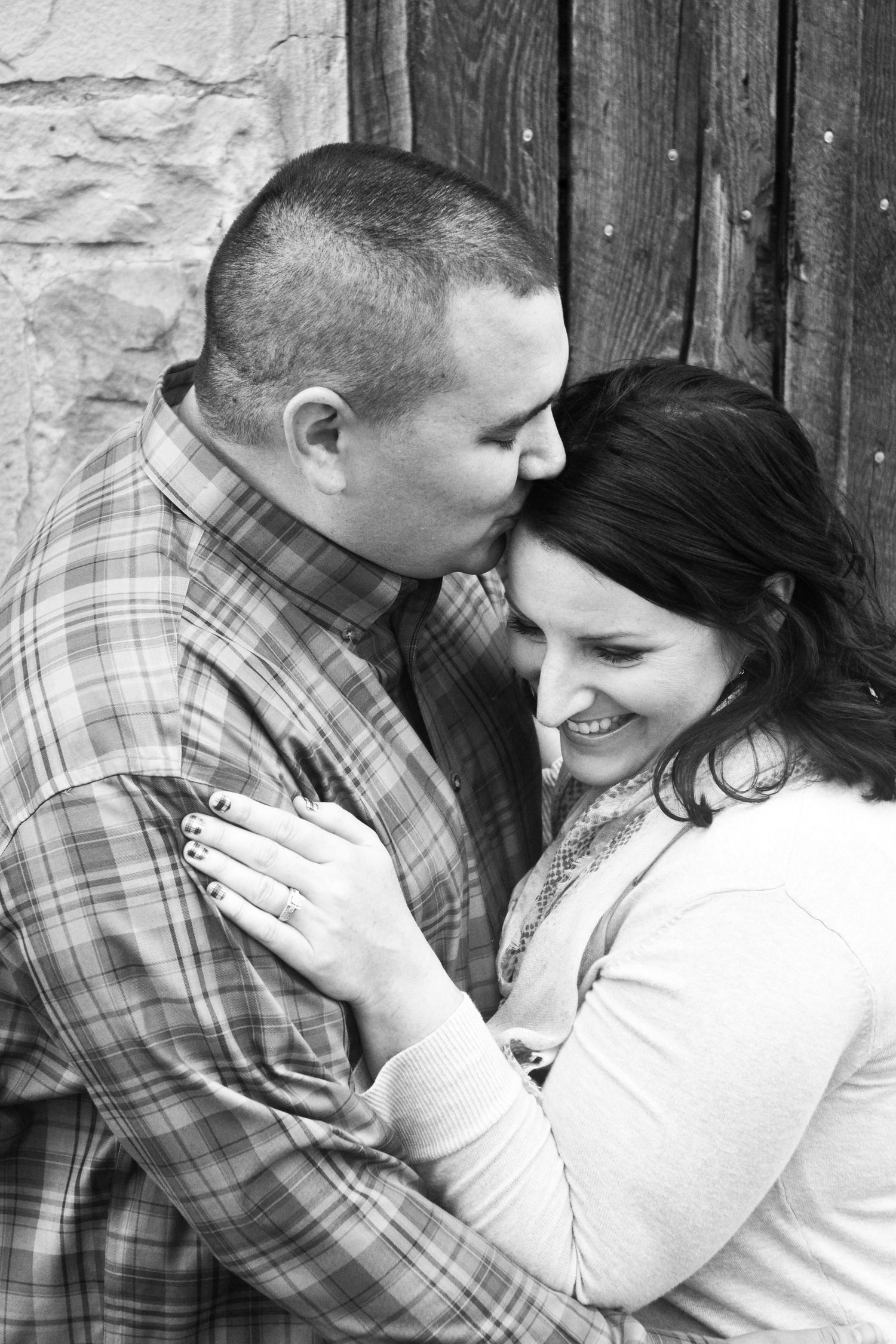Black and white photo of a couple embracing near a rustic wooden door during an engagement session.