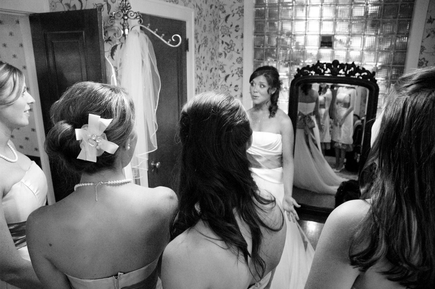 Bride and bridesmaids gathered around a mirror in a vintage floral room during wedding preparations.