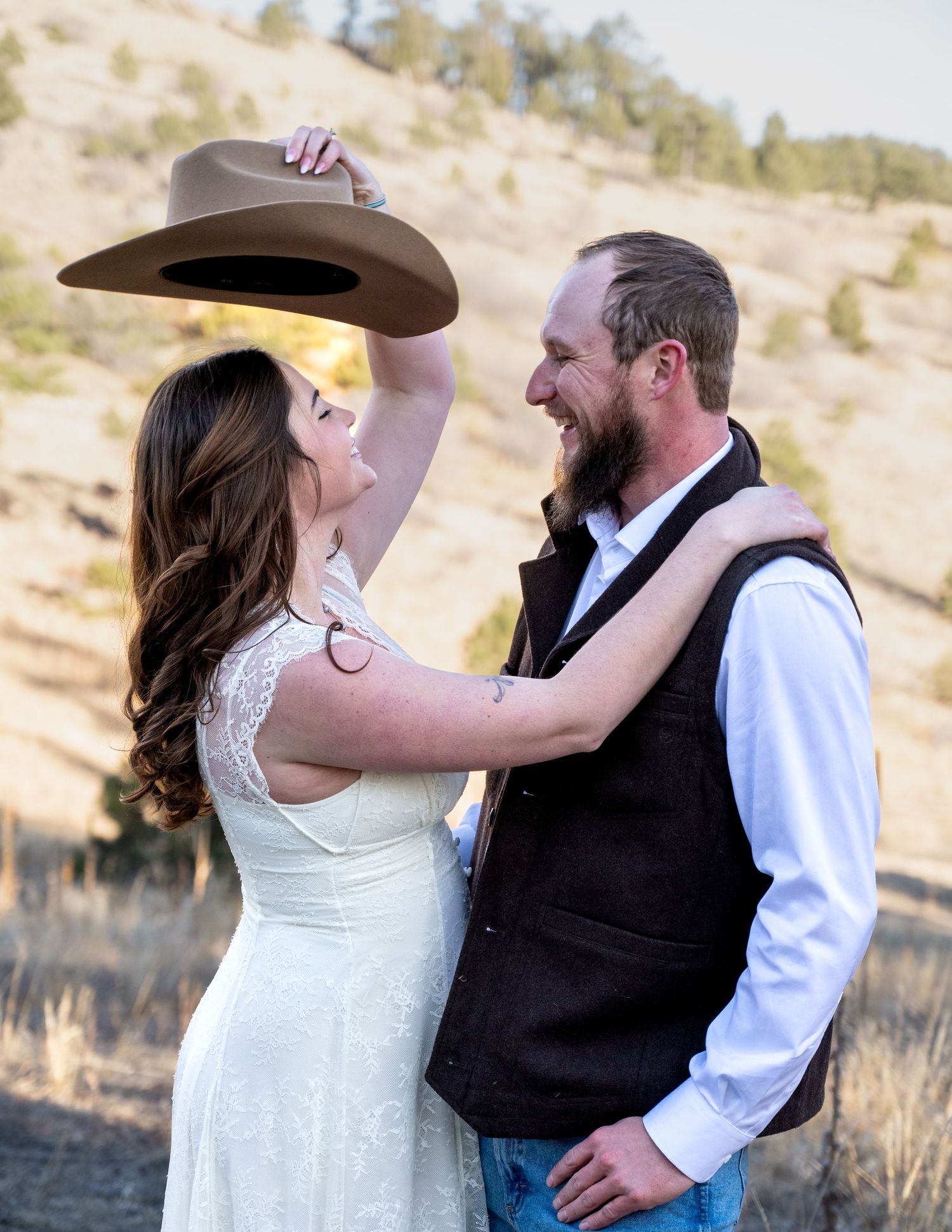 Bride playfully holding cowboy hat over bearded groom while smiling in a rustic outdoor setting.