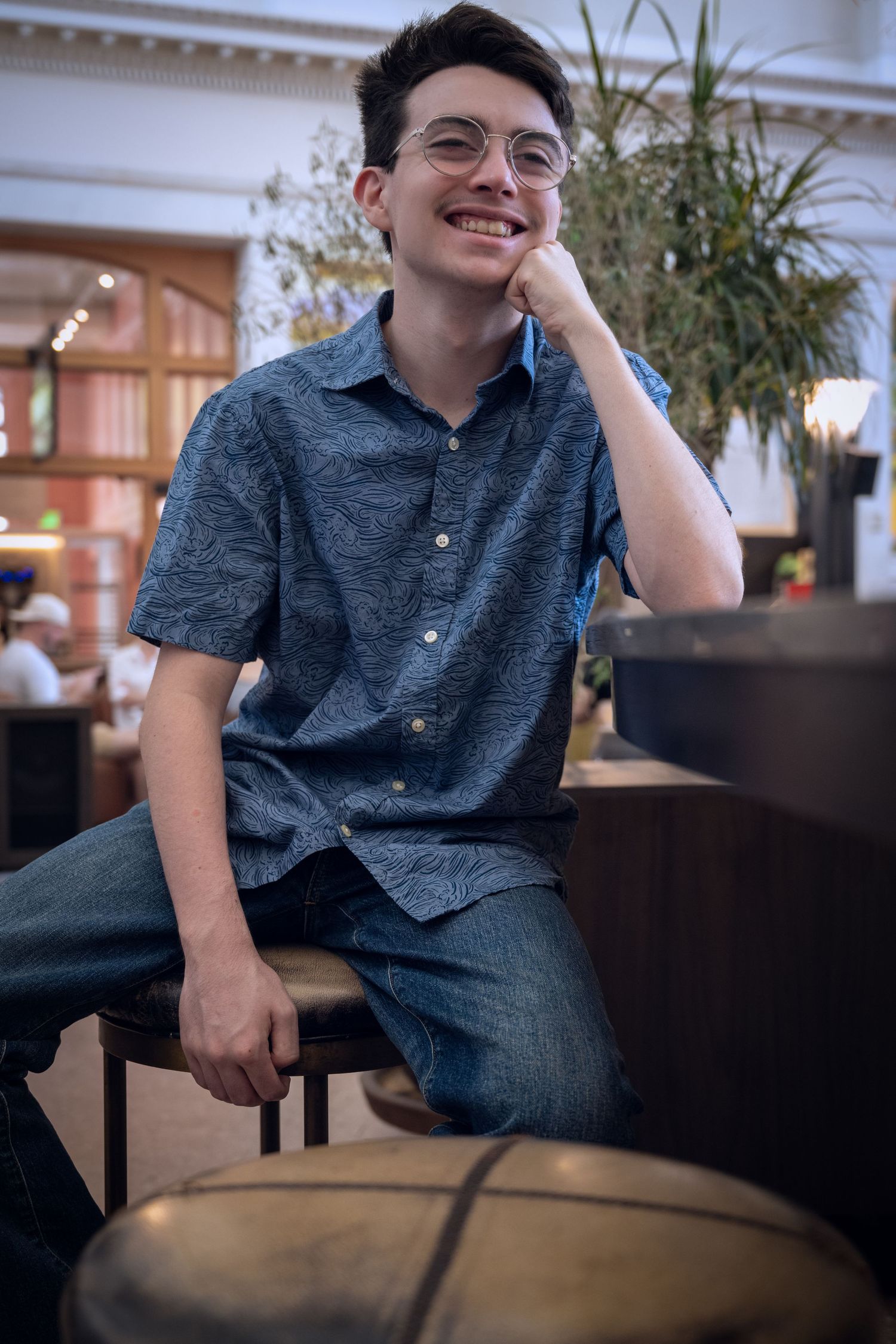 Smiling young man with glasses sitting on a stool inside a bright indoor space.