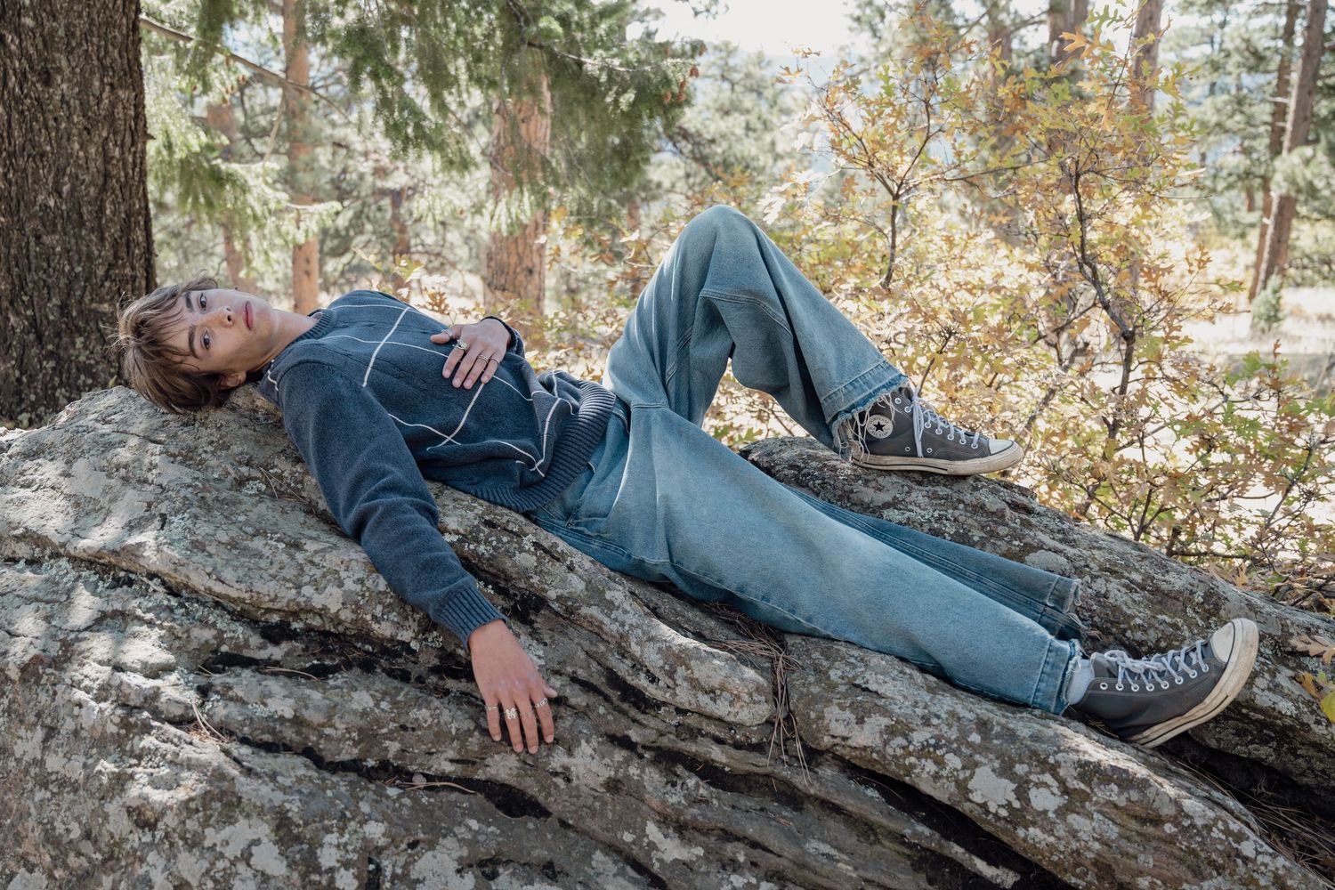 Young person lying peacefully on large rock in forest wearing jeans and navy hoodie.