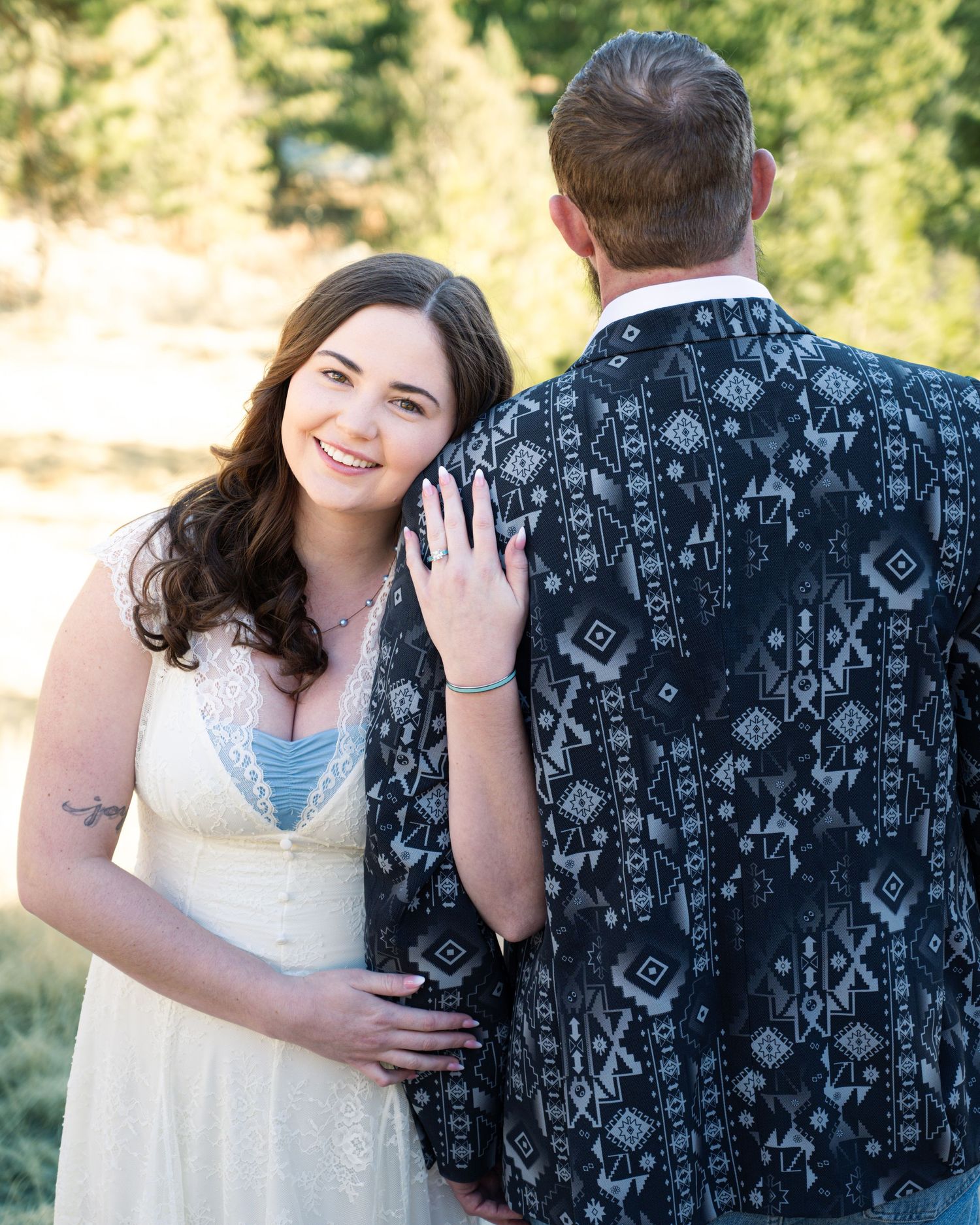 Smiling bride in lace dress placing hand on groom's patterned jacket during outdoor photo session.