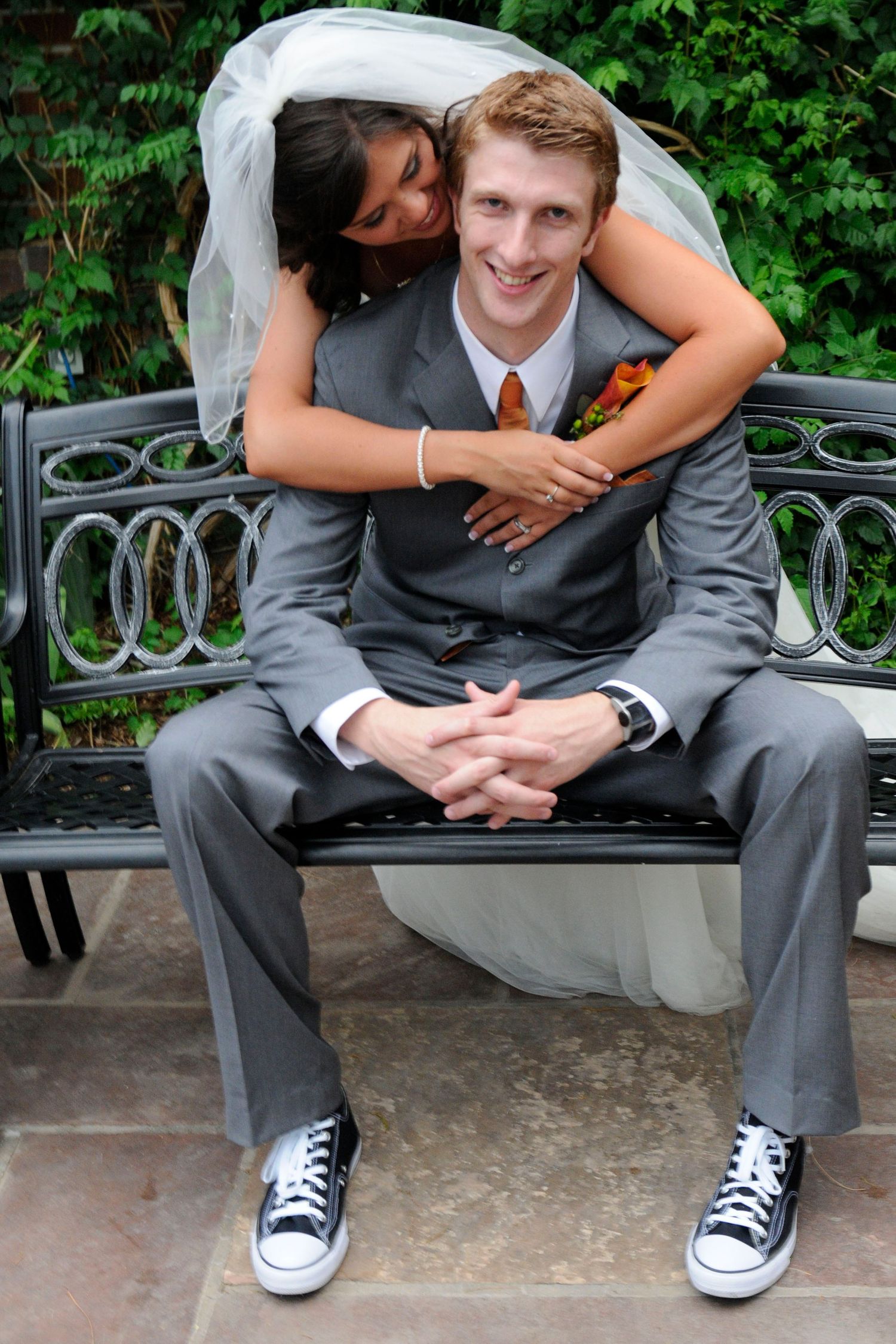 Smiling groom in gray suit and Converse sneakers sits on bench as bride embraces him from behind.