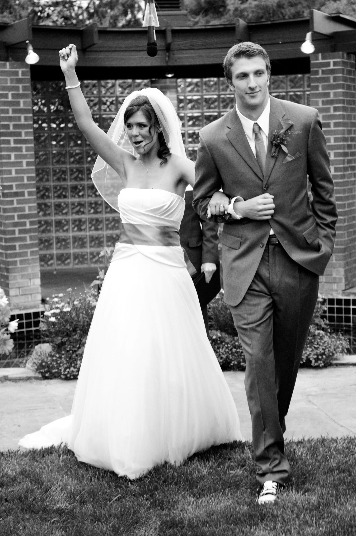 Black and white photo of a joyful bride raising her fist while walking with her groom after their wedding ceremony.