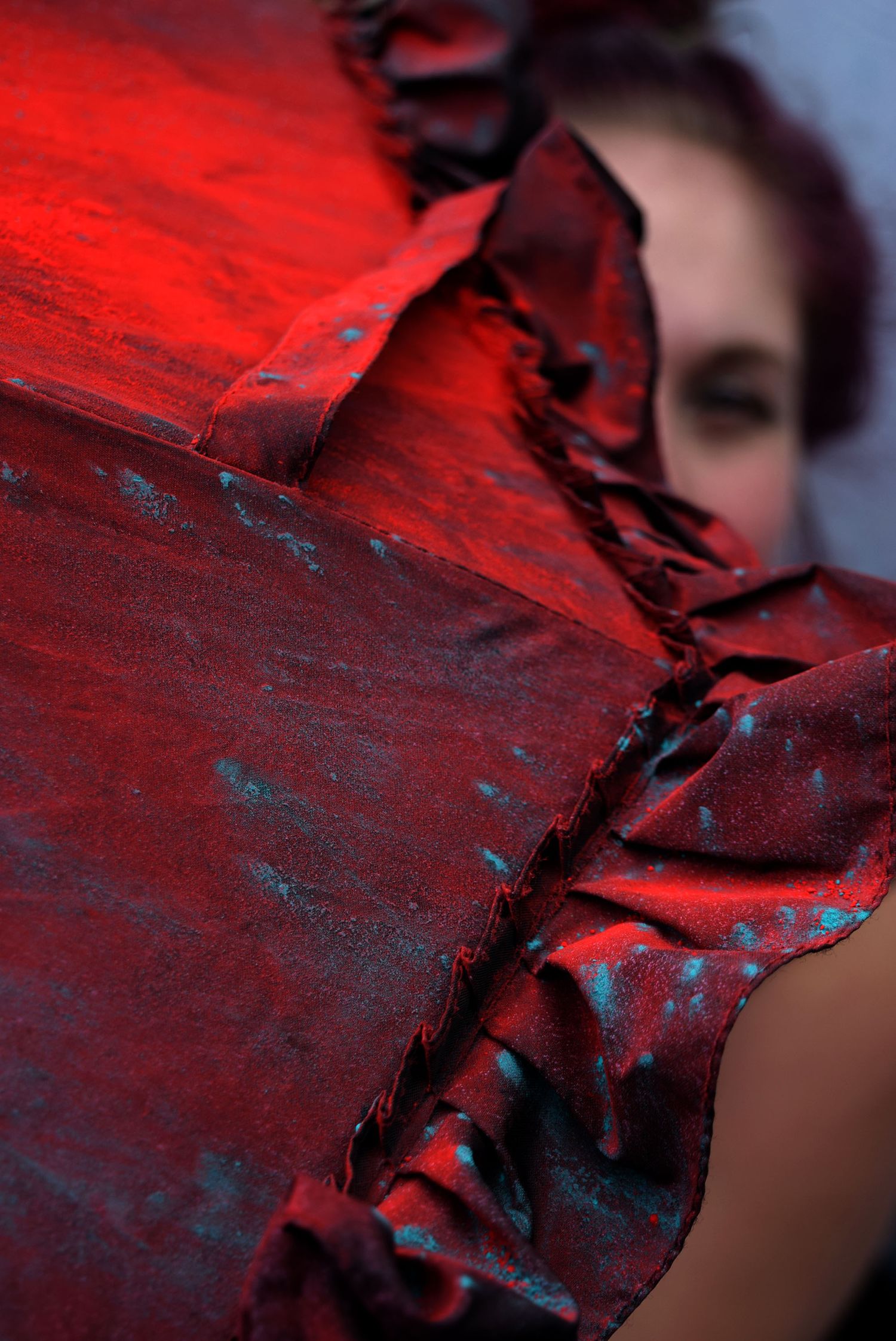 Close-up of a textured red ruffled fabric dress with teal accents, worn by a blurred woman in the background.