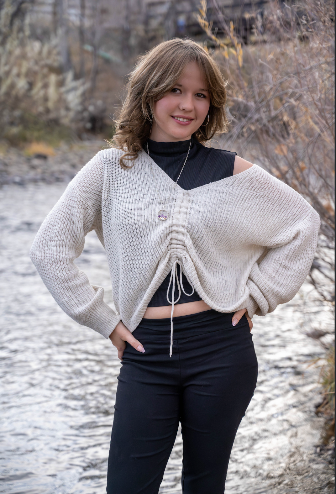 Teen girl in cream knit sweater and black outfit posing by a rocky stream.