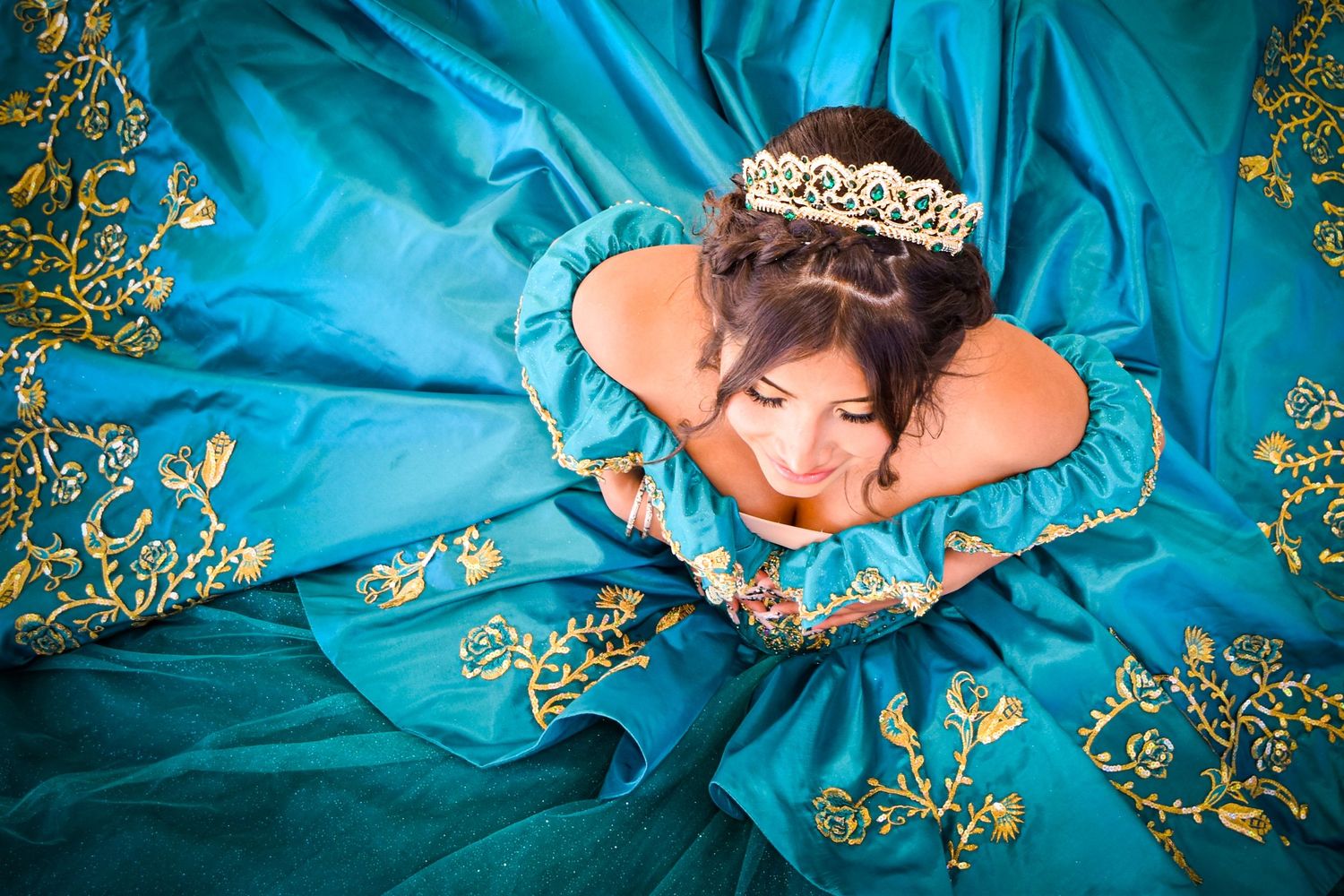 Young woman in teal ball gown with gold embroidery and tiara poses gracefully for quinceañera portrait.