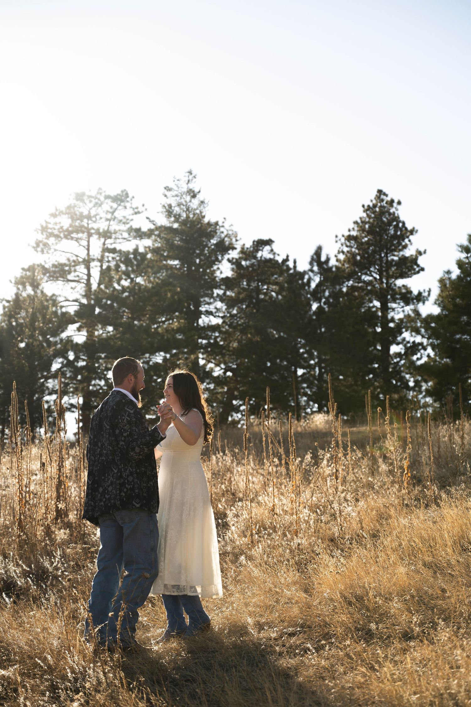 Couple dancing in a sunlit golden meadow surrounded by tall pine trees at sunset.